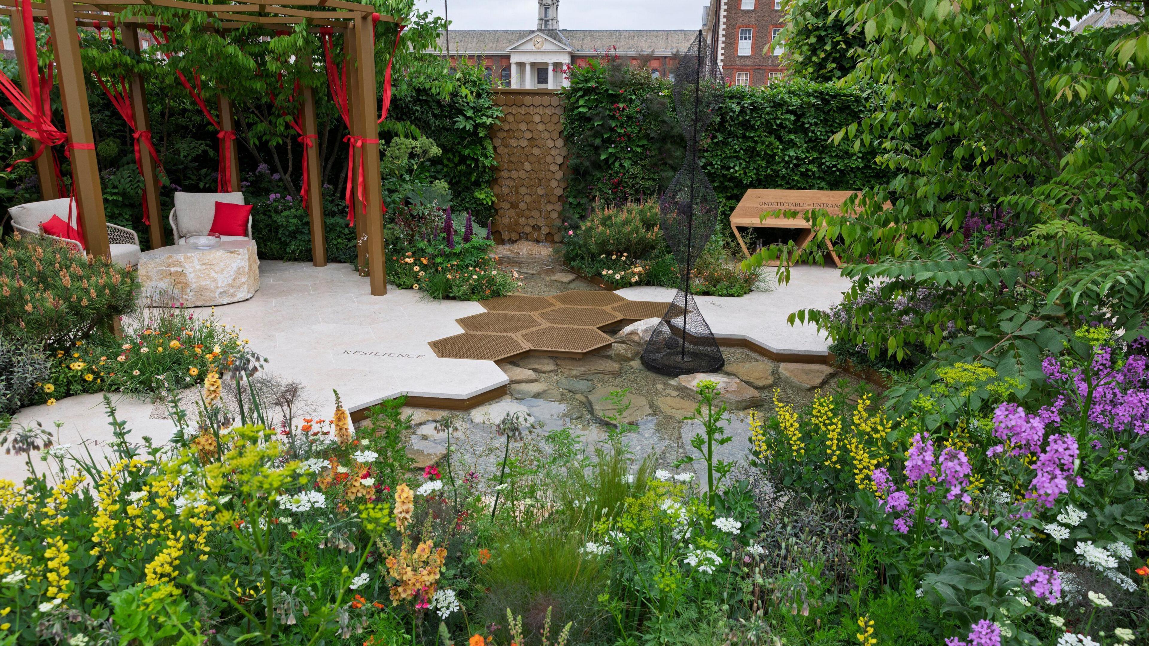 A picturesque garden seen with colourful flowers blooming. There is a seated area in the corner with red ribbons hanging from it.