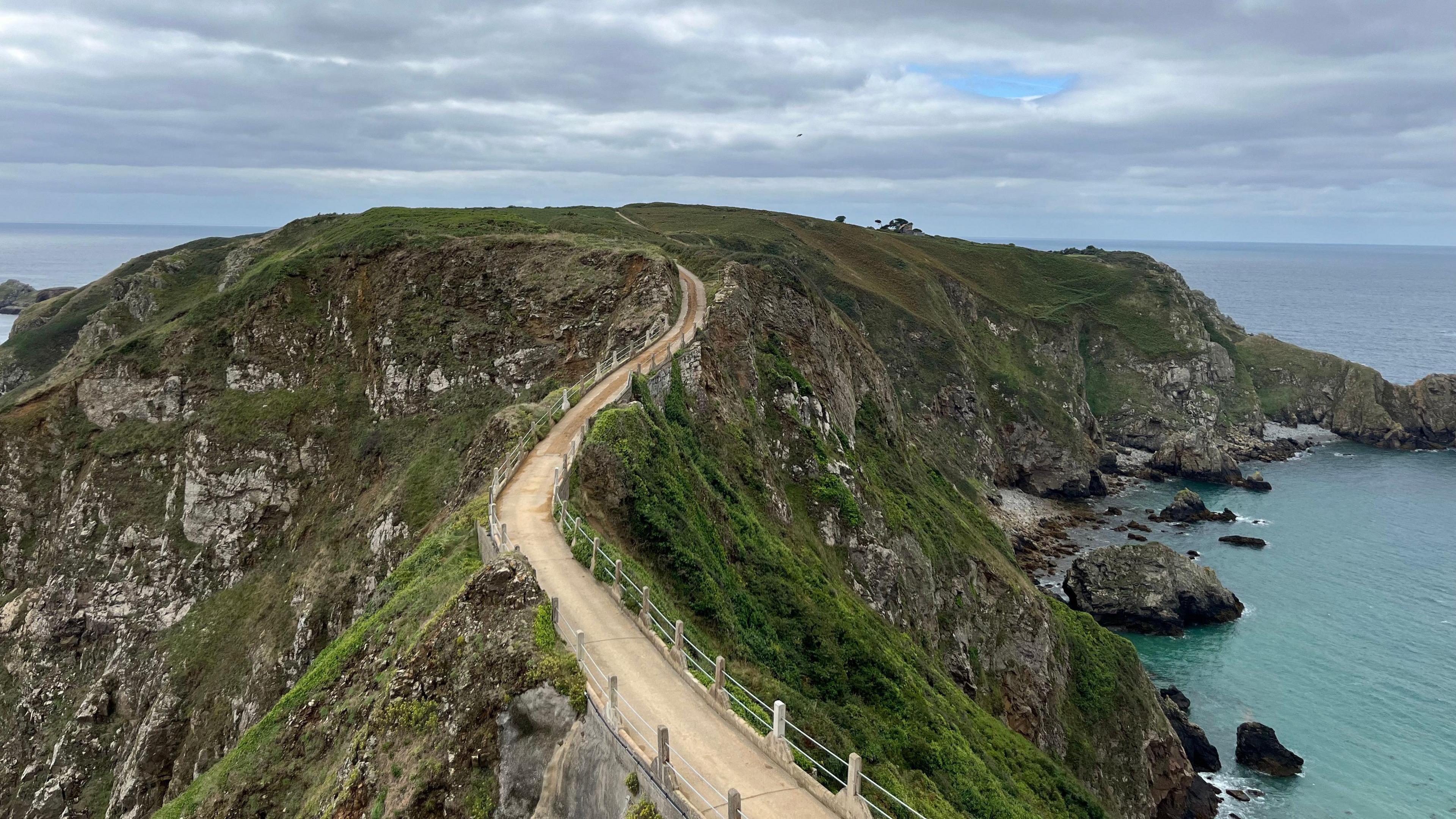 Aerial view of La Coupée in Sark showing a yellow road winding up a green mountainous hill, with the sea to the right