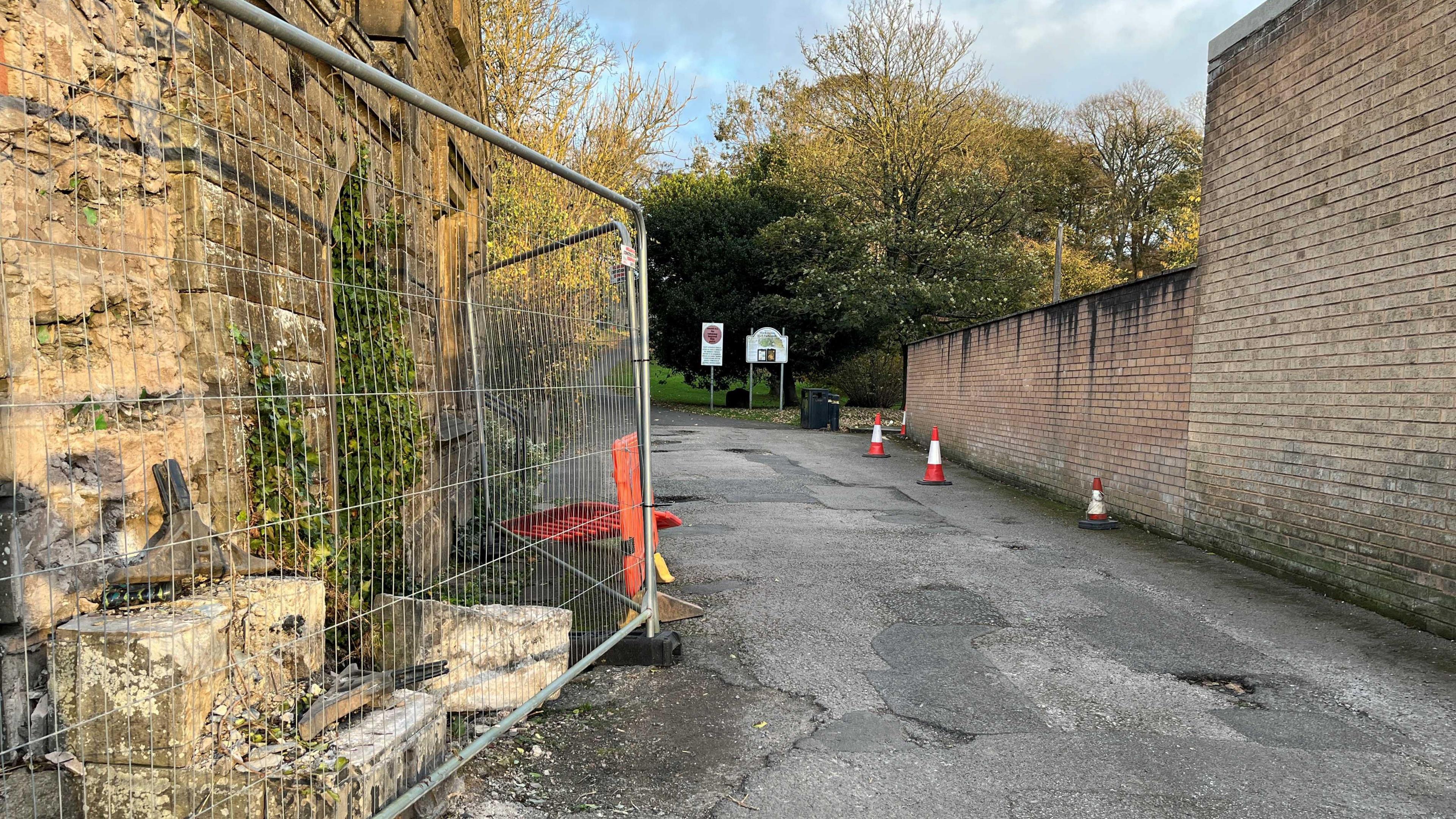 Heras fencing surrounds the gable end of the Hall Park Lodge property where the damage has been caused. The park, with grass and trees is visible in the background.