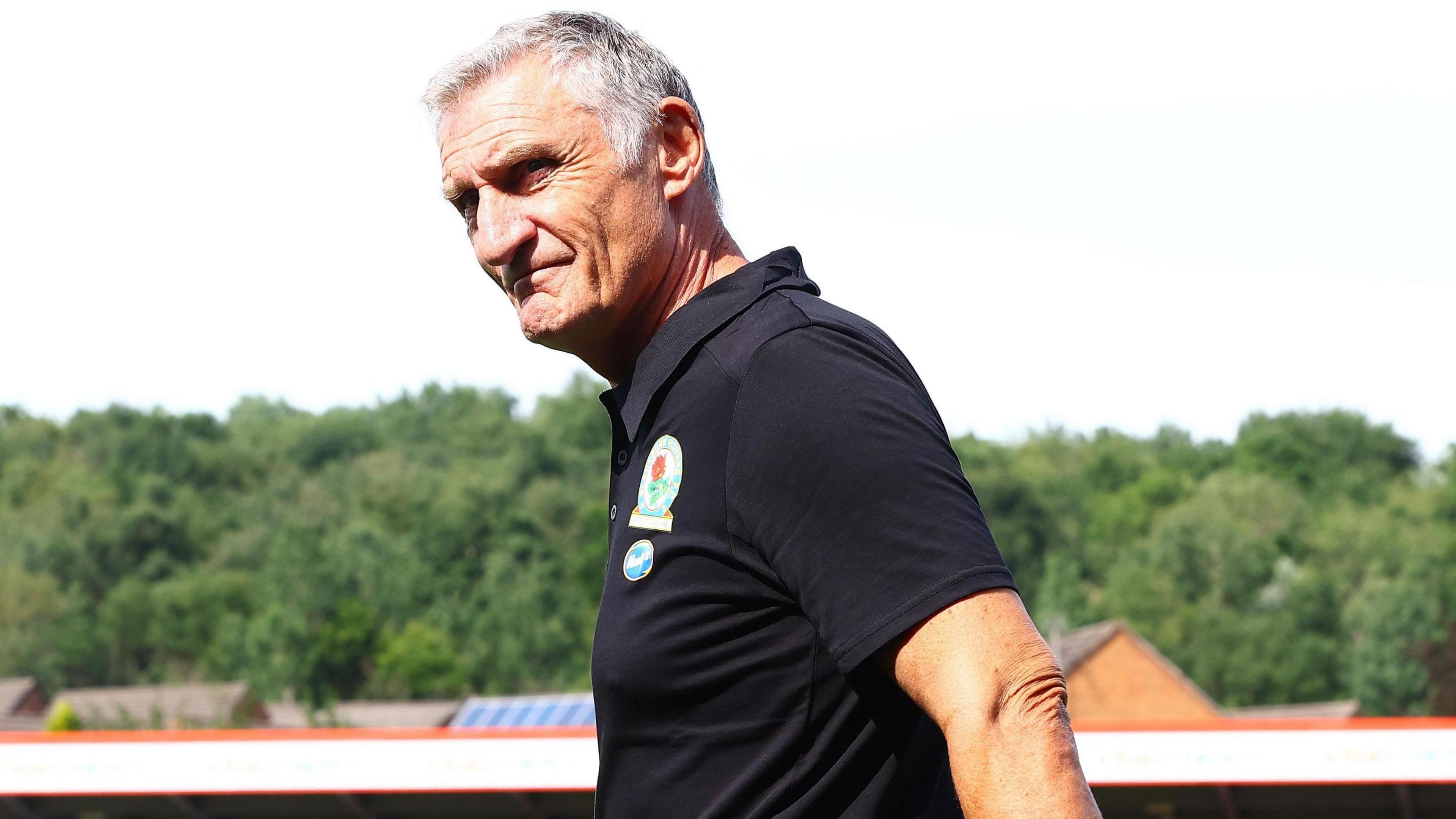Blackburn Rovers manager Tony Mowbray walks acrToss the pitch with a stand and trees at Accrington Stanley's Wham Stadium in the background.