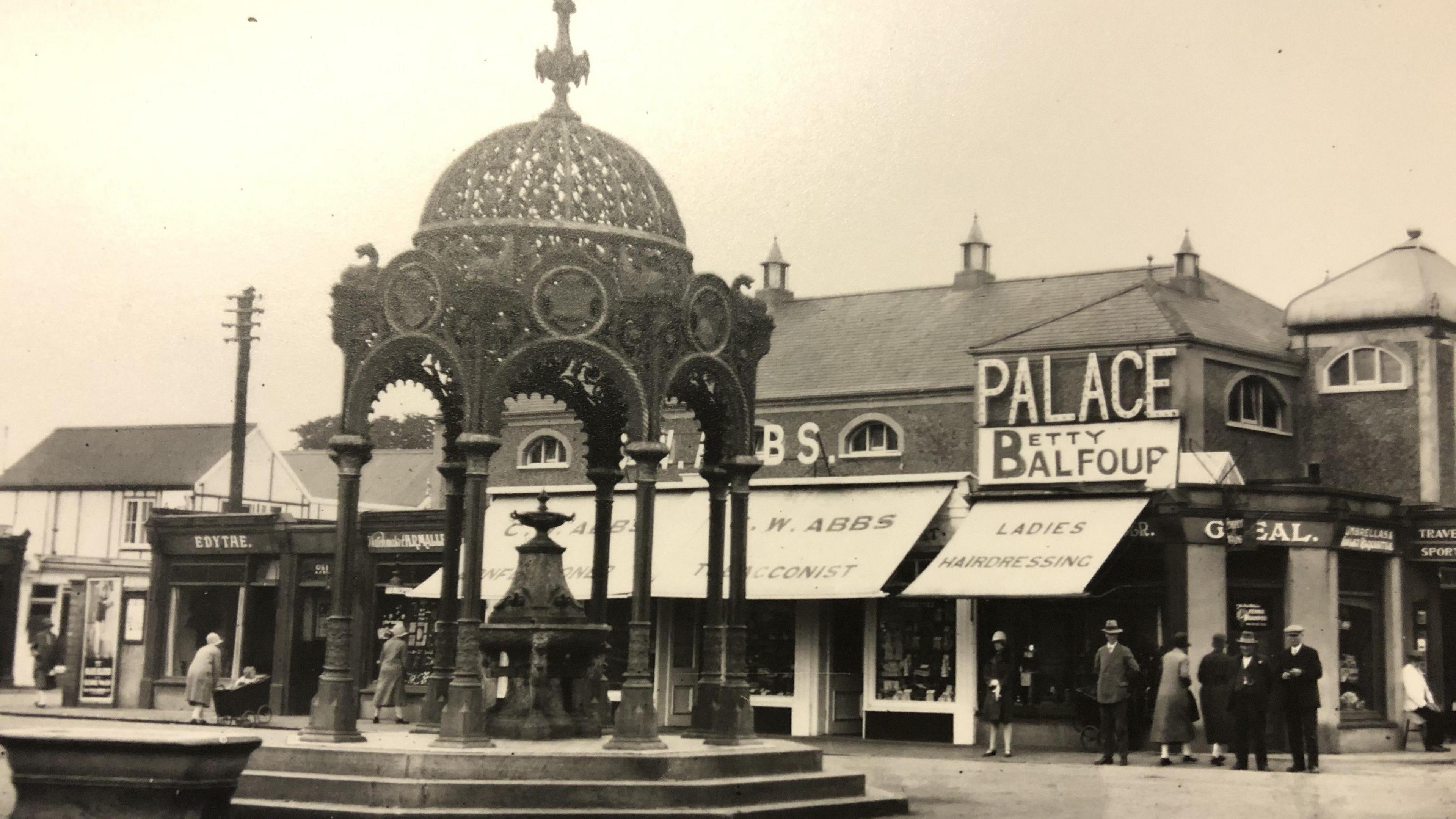 Black and white picture of the Coronation Fountain from 1929, complete with water drinking fountain in the centre. Smartly dressed men and women can be seen near shops in the background, wearing hats suits and coats.