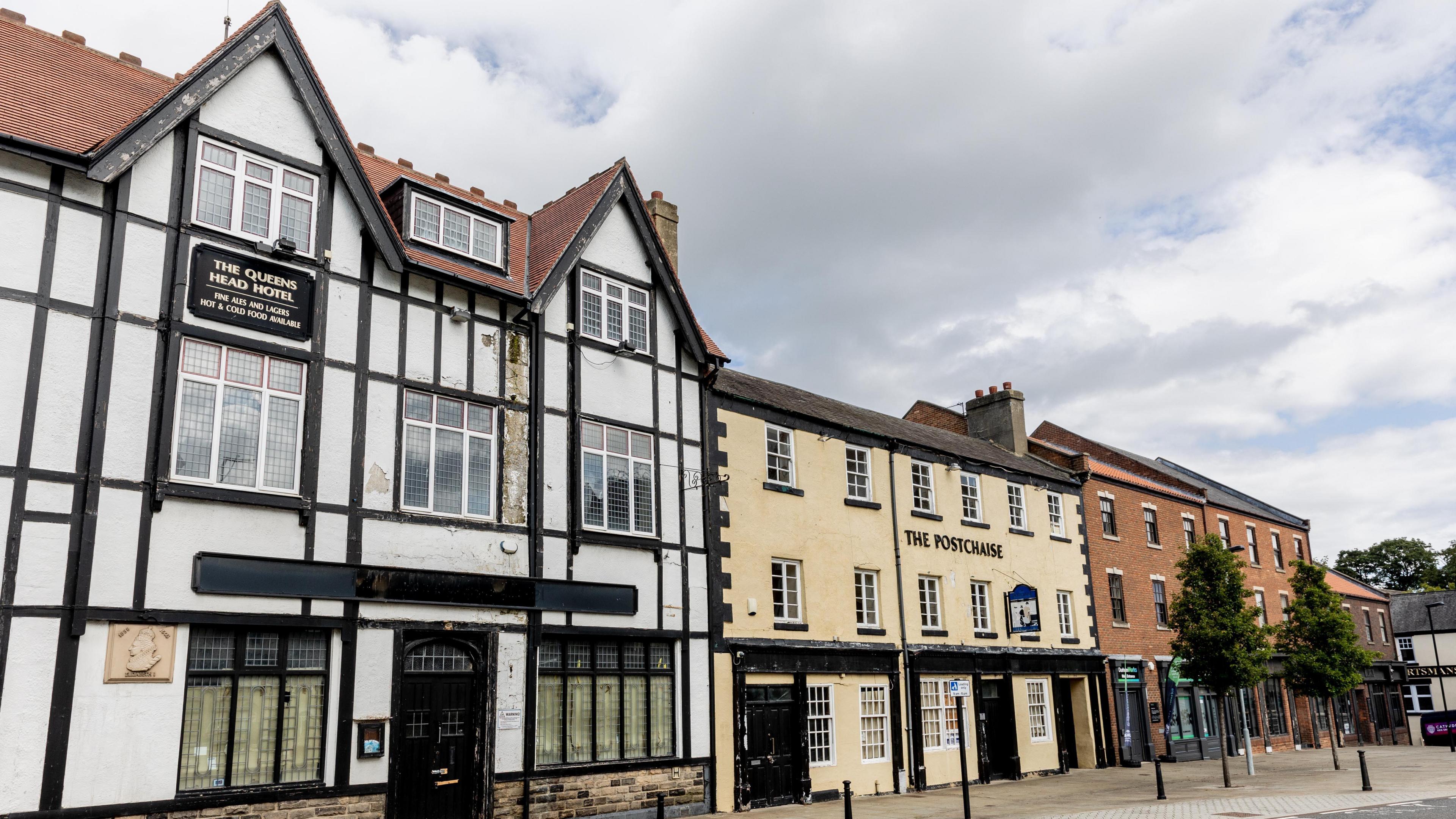 The Queens Head, Postchaise and Monaco buildings are three-storey buildings that stand side-by-side. The Queens Head has a black and white timber design on the exterior. The Postchaise is painted beige and the Monaco is a red-brick building with black doors.