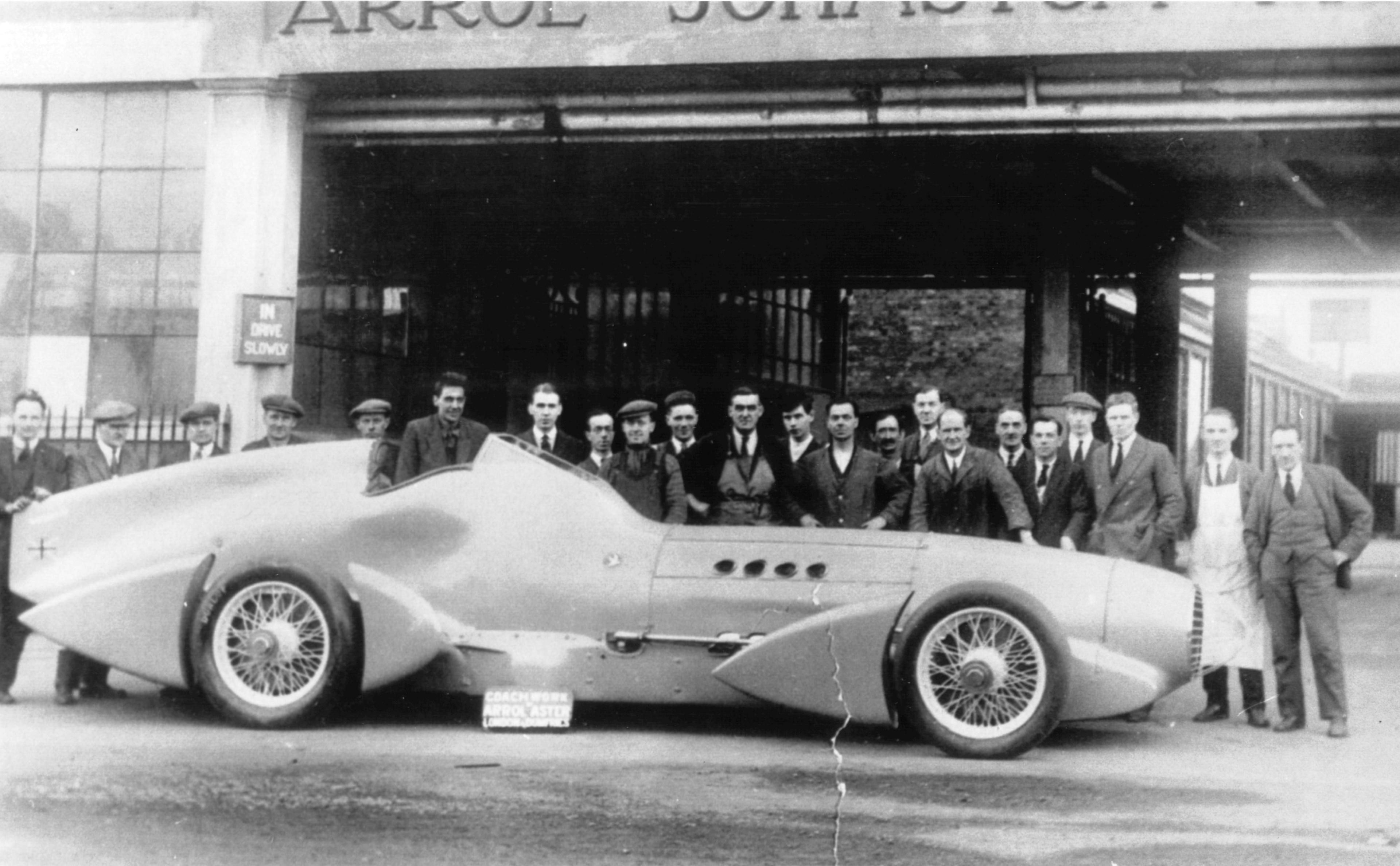 A group of workers - many in bunnets - line up behind a massive racing car from the 1930s