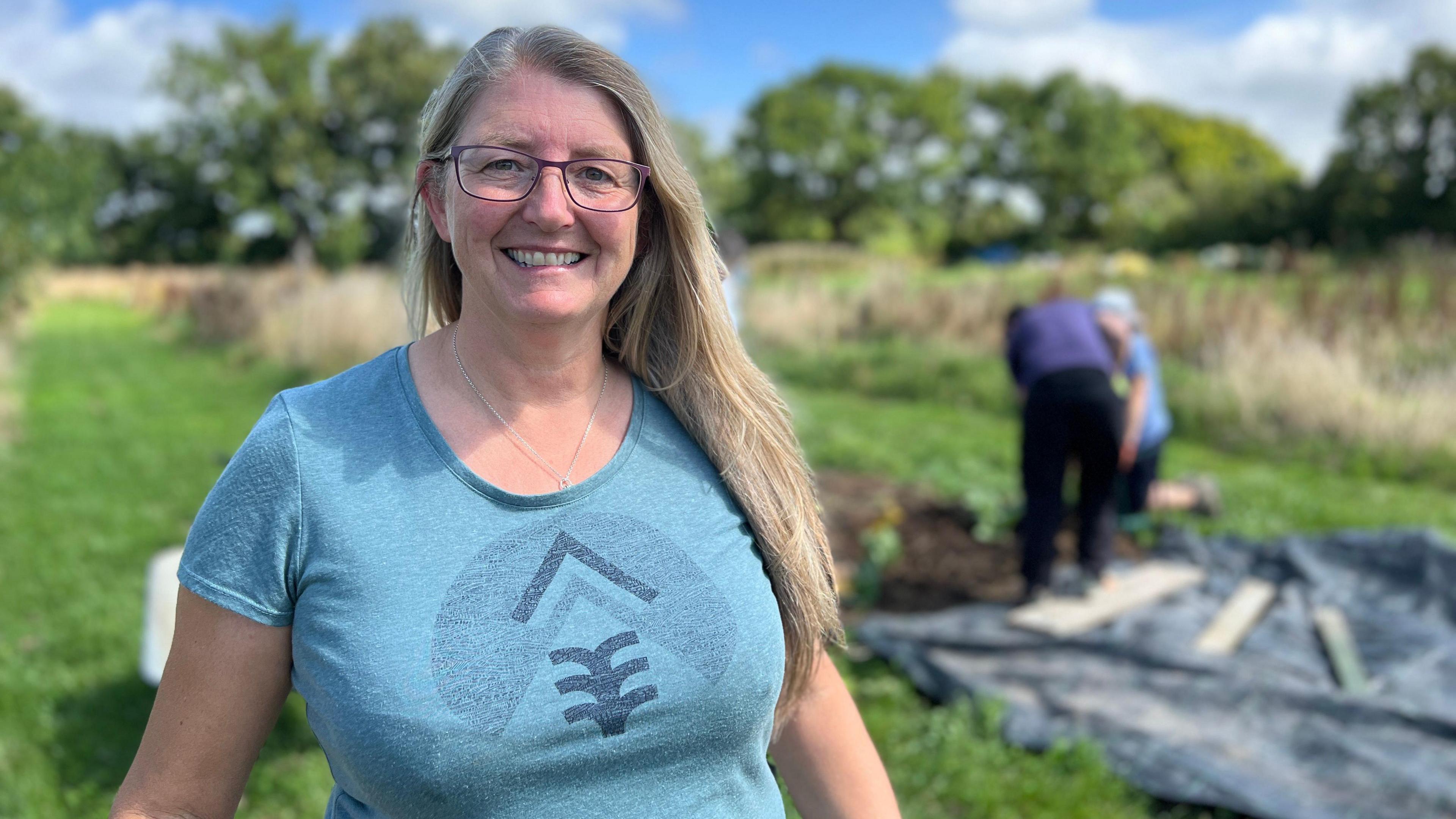 Sheena smiles at the camera as people work and dig in a field beind her