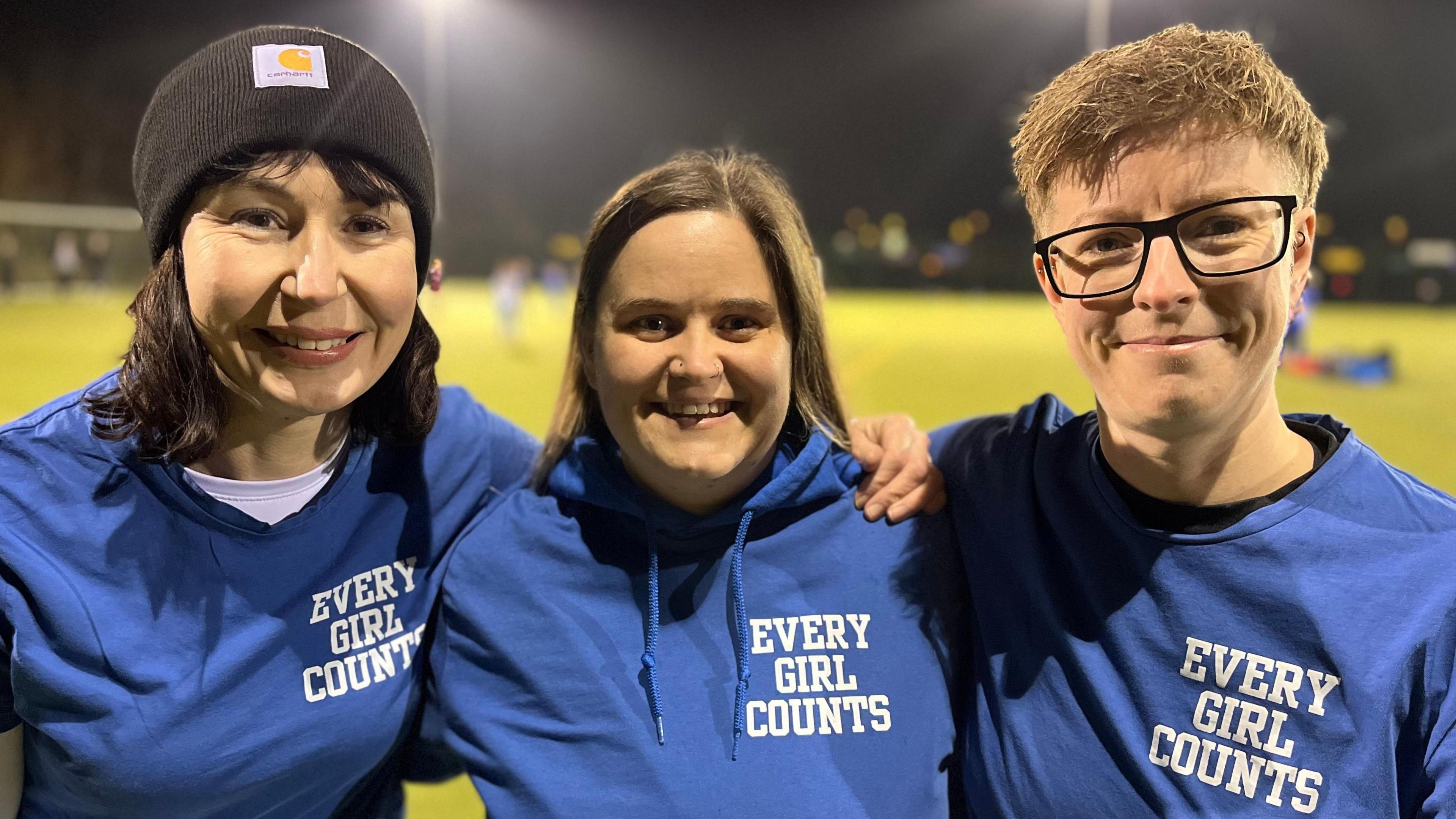 Three women wearing t-shirts saying "Every Girl Counts" are looking at the camera.
There is a floodlit football pitch behind them.