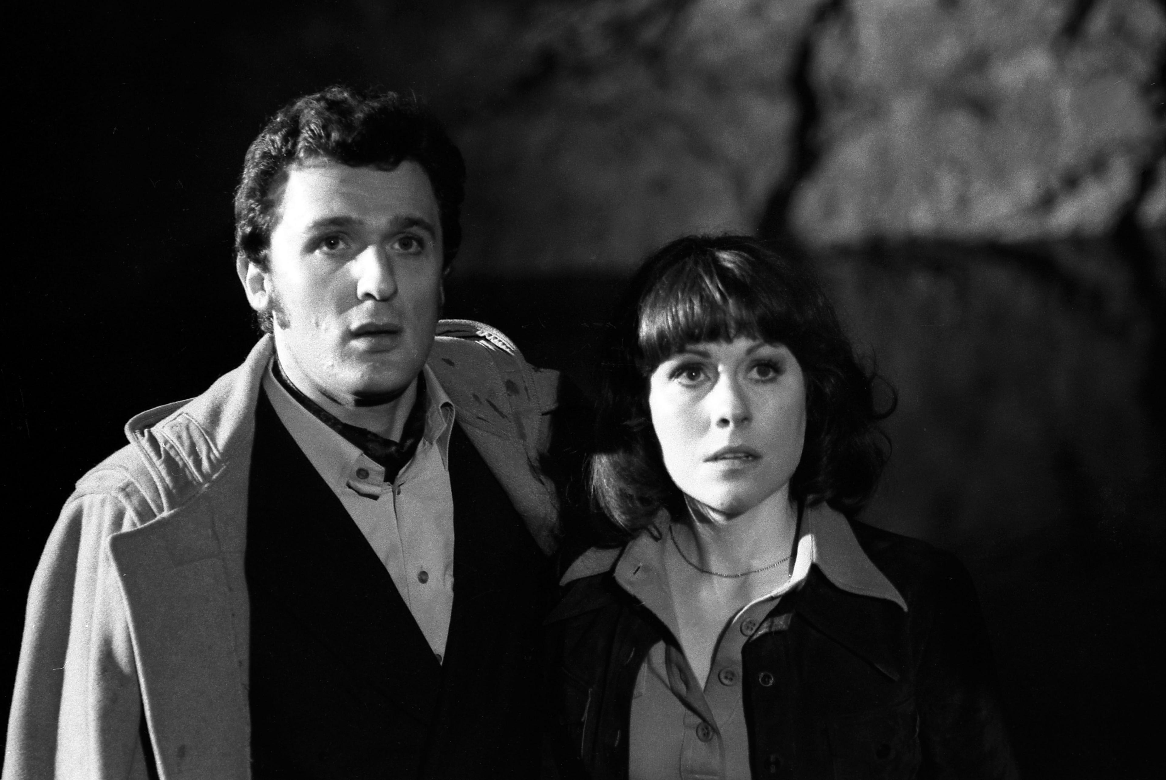 Black and white photo of Harry, who has short dark hair and is wearing a duffle coat over an open necked shirt and dark blazer, standing next to Sarah, who has dark, shoulder length hair and also wears an open necked shirt and dark jacket. They are standing in a cave with the light casting shadows on the wall behind them.