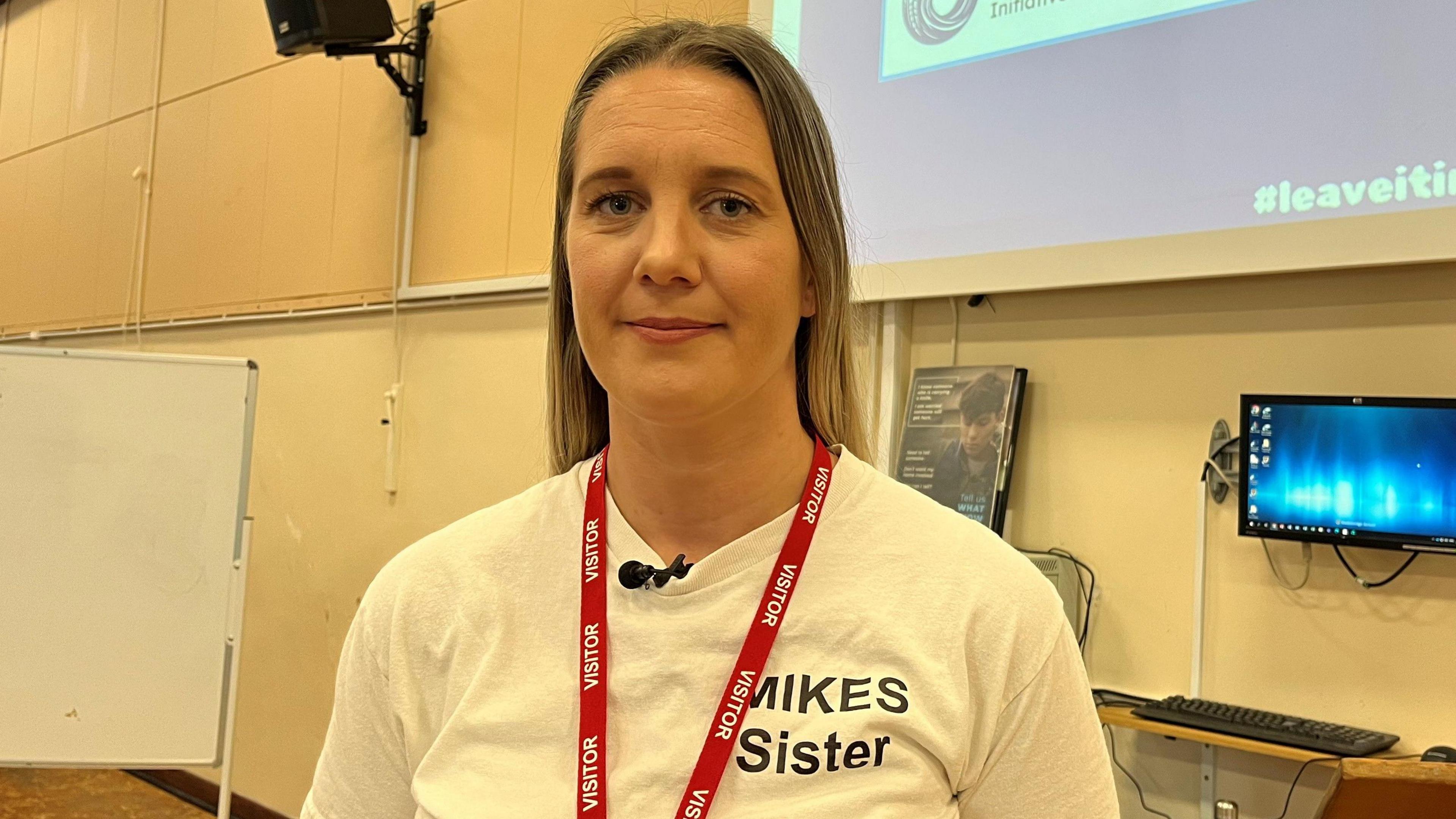 A woman in a white t-shirt stands in front of a class of year 10 students in a school hall. Behind her is a picture of her brother who was stabbed to death.