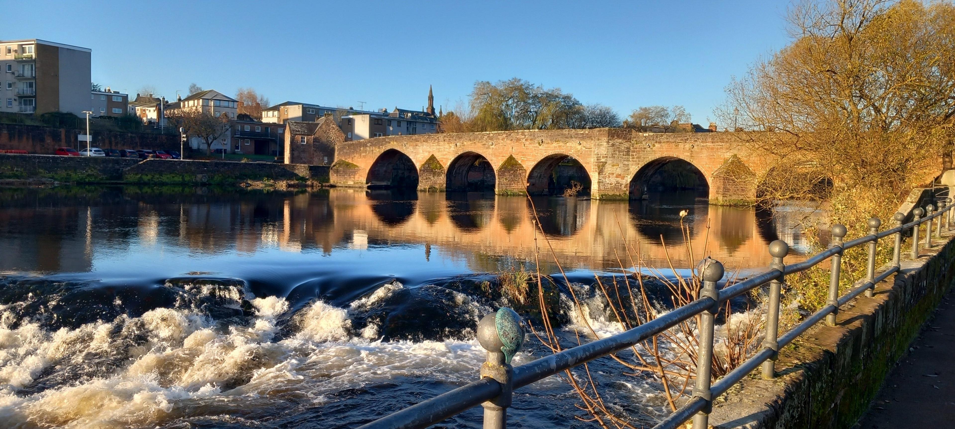 An old sandstone bridge has its arches reflected in the river below with some blocks of flats in the background