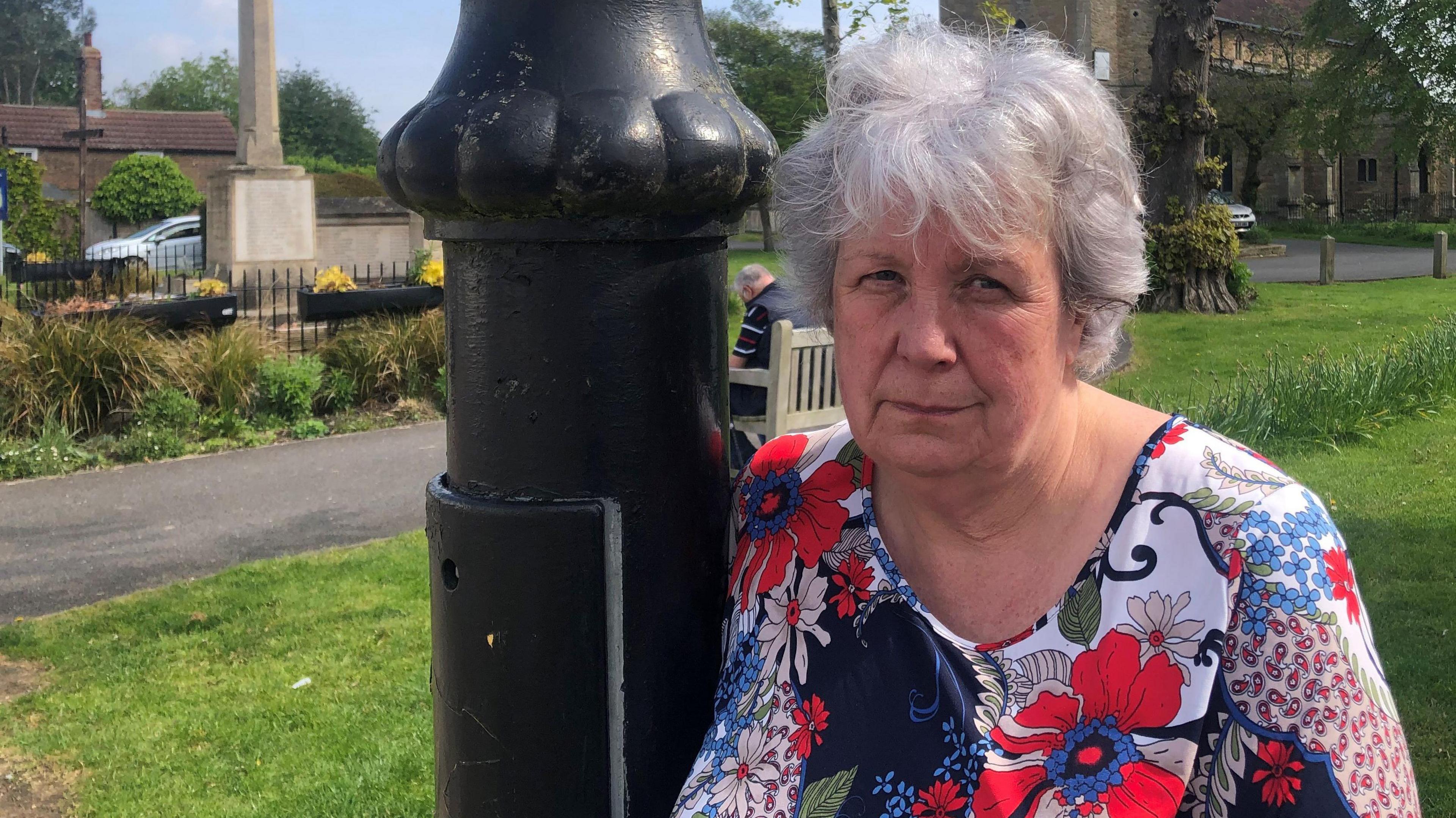 Sue Unwin has short grey hair with a fringe over her forehead. She is wearing a floral design dress with a low cut round neck and is leaning against one of the heritage lamp-posts. Behind is a church and a war memorial, with greenery seen too.