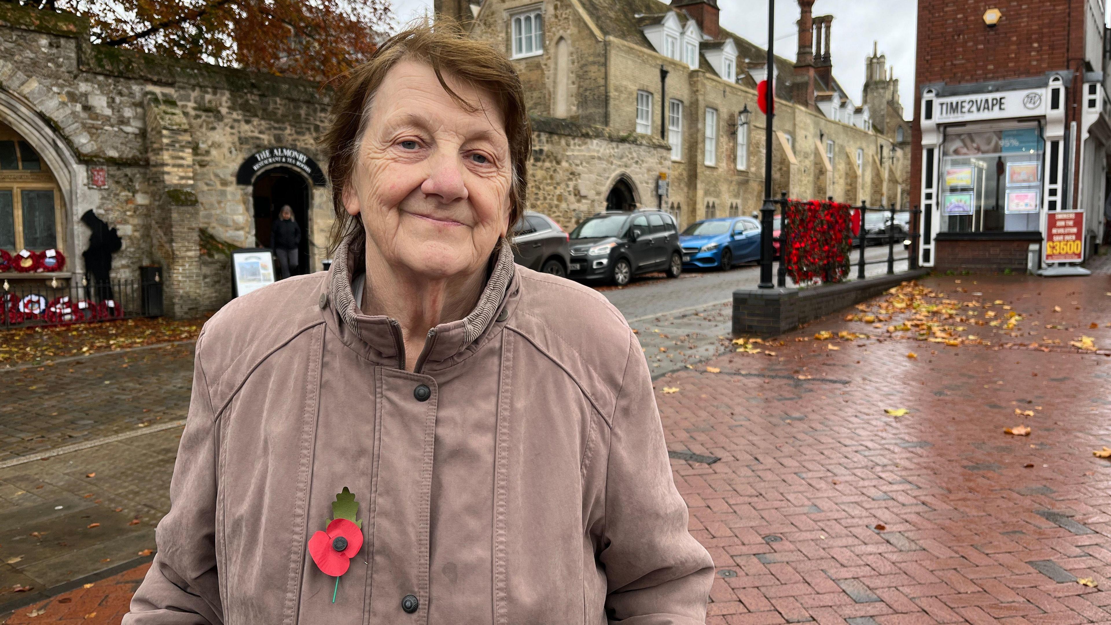 Patricia Canham is standing to the left of the photo and smiling at the camera. She is wearing a light-coloured jacket with a red paper poppy affixed on the left-hand side. Behind her is a sandstone-coloured wall with a war memorial in an alcove including the figure of a black metal solider with his head bowed and several poppy wreaths. There are also poppies adorning a nearby railing and one attached to a lamppost. 
