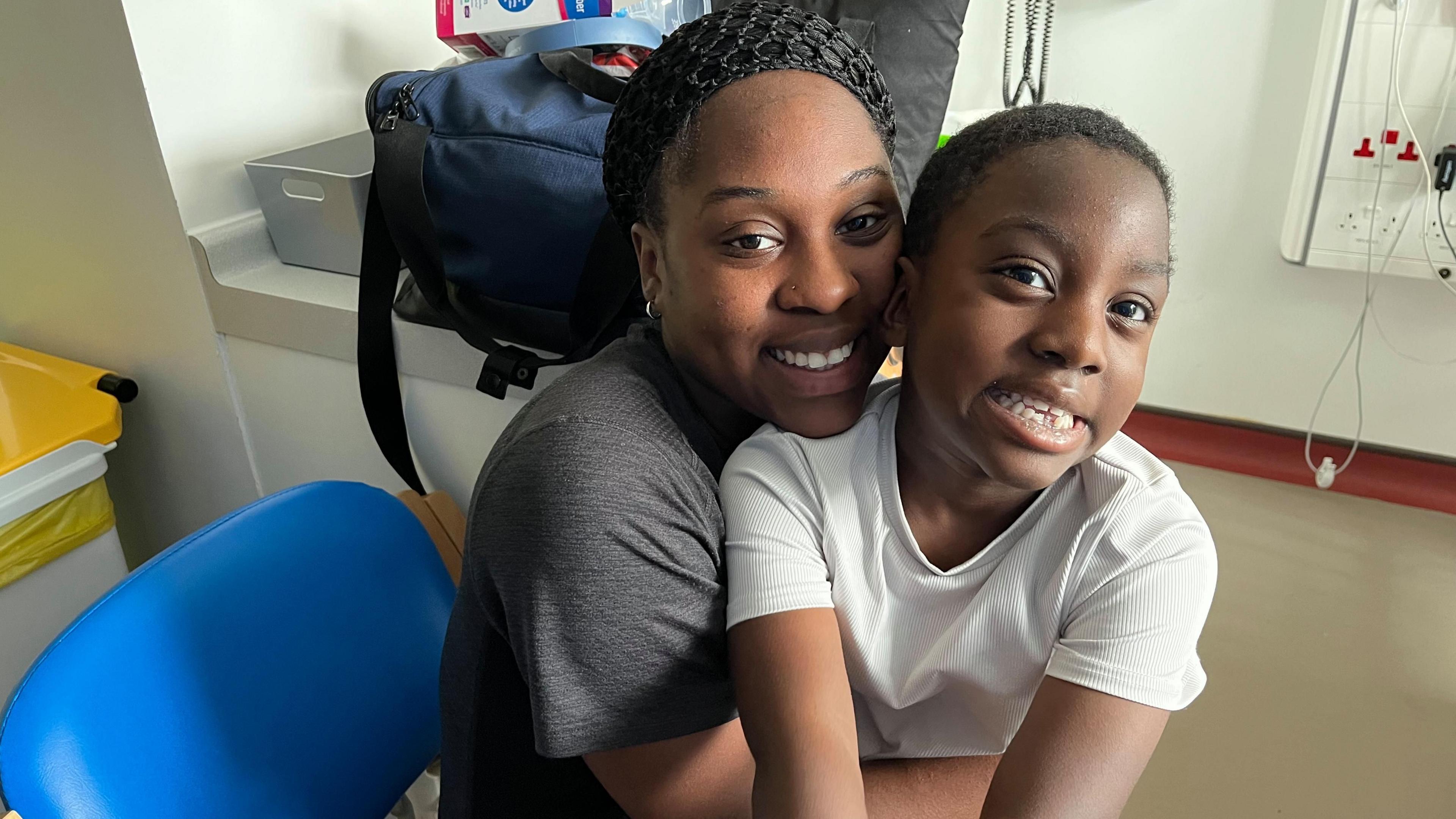 Woman with short dark hair in black top with son with short dark hair in white top sitting on a blue chair. They are both smiling. There is a large blue bag on a shelf behind them.