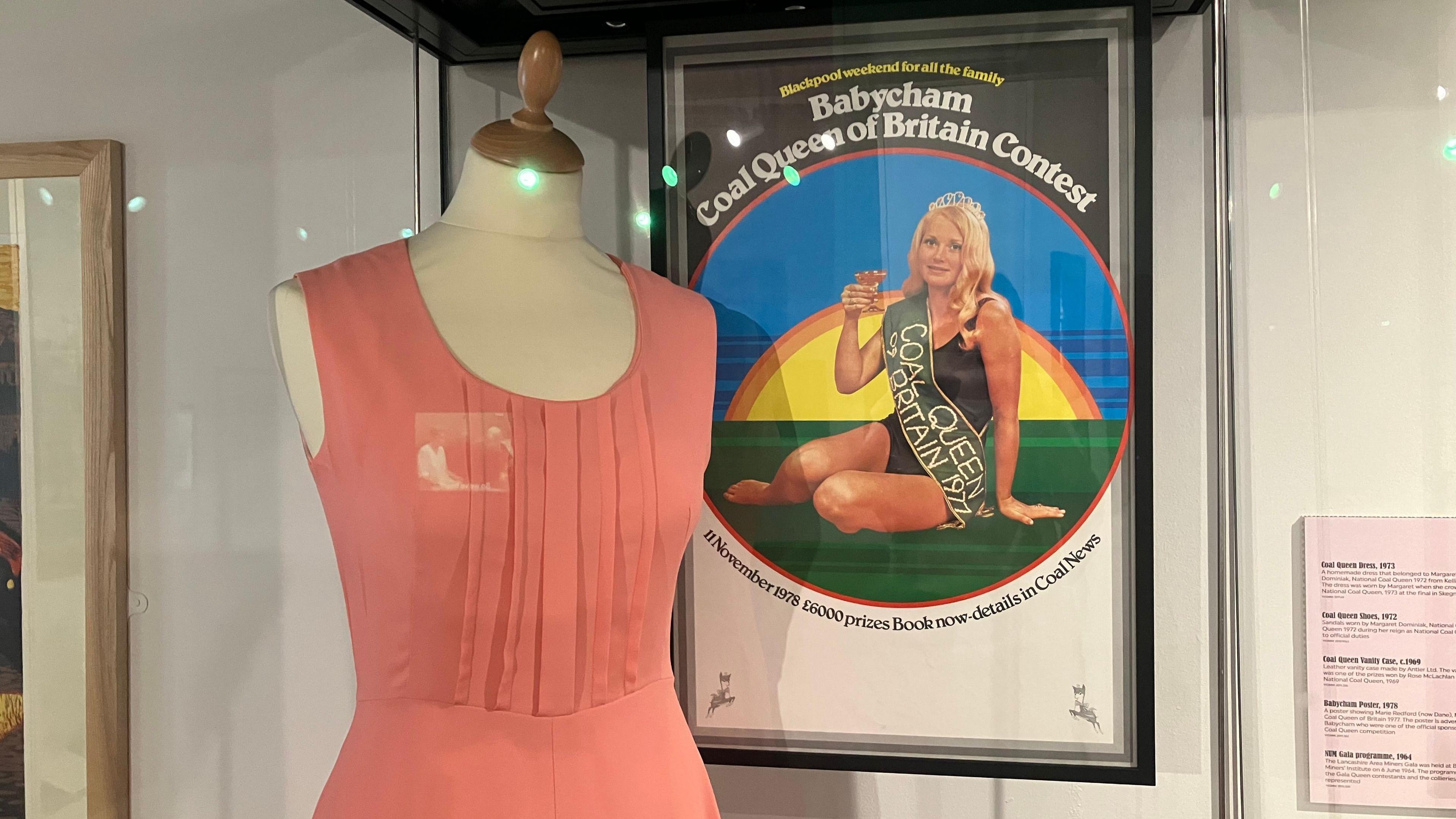 A manequin wears a peach dress. Behind it a poster of a "coal queen" hangs. The poster depicts a blonde woman with a coal queen sash in front of a setting sun.