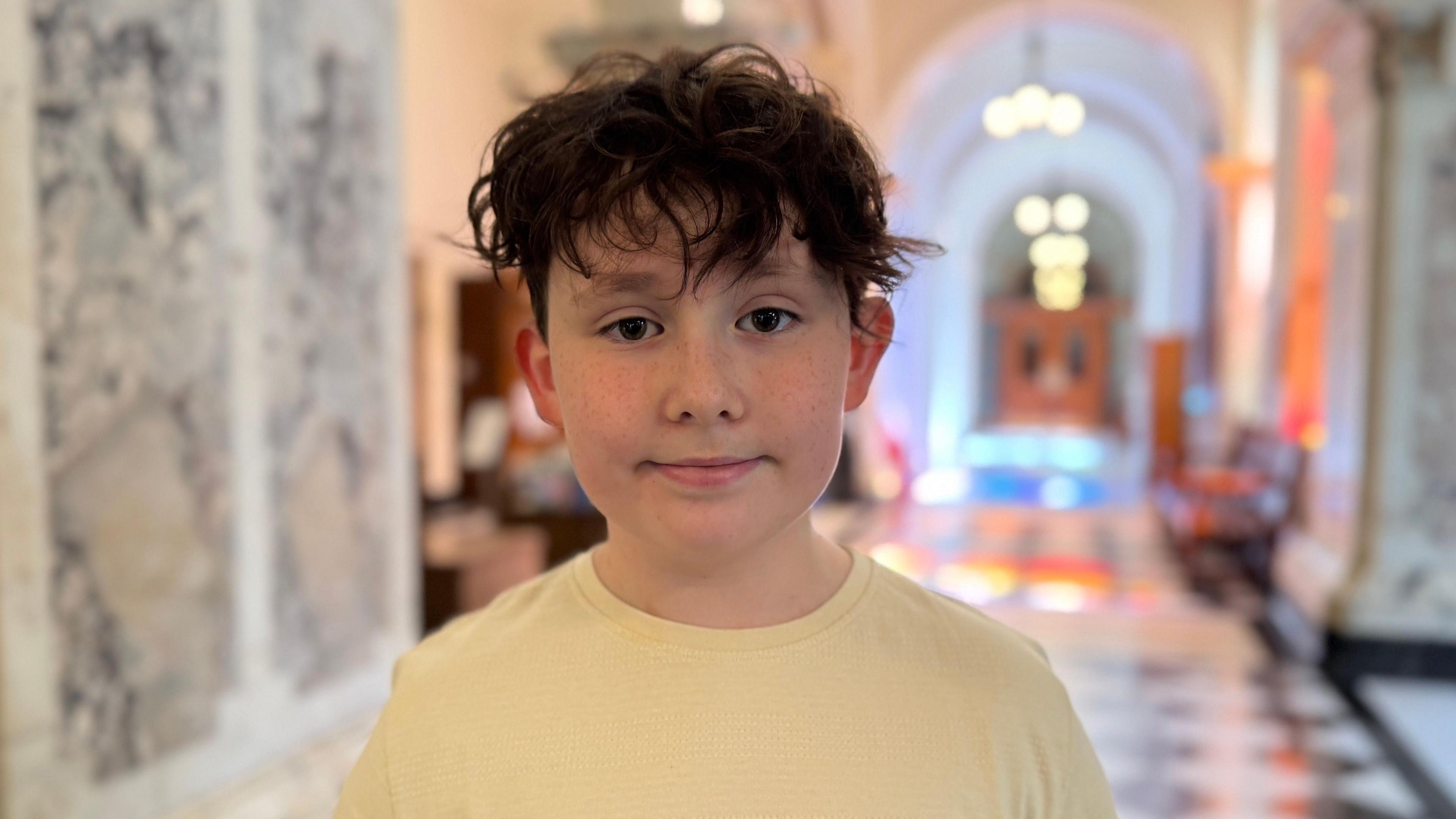 Jack has long brown hair and is smiling at the camera. He is wearing a pastel yellow t-shirt. He is stood in a hall with marble walls and floors but the background is blurred.