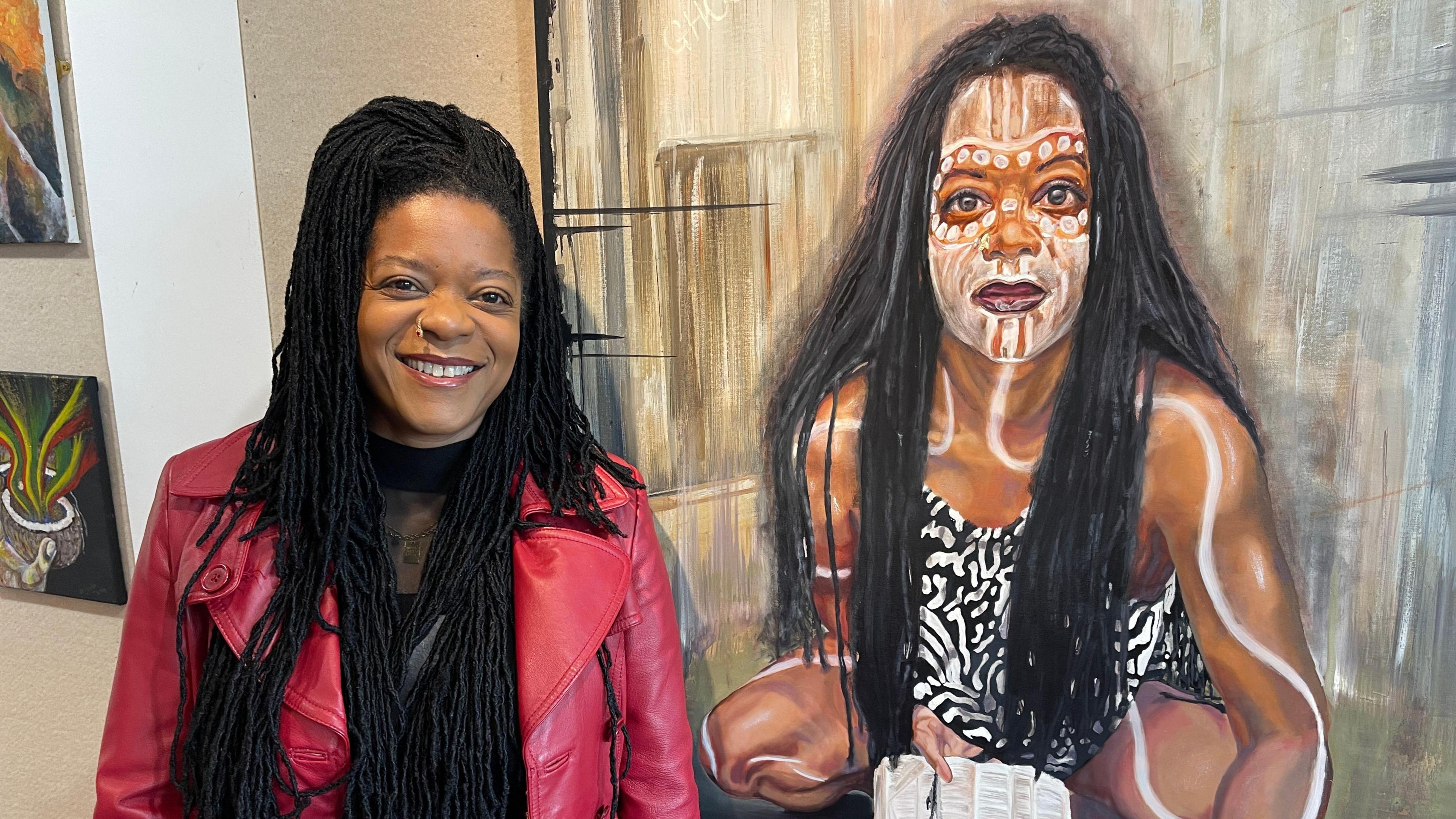 Artist Esme Layne, who works in oil on canvas, is stood beside her self-portrait, which she says is an exploration of her identity. She is smiling, has long long dark hair, and is wearing a pink jacket and black top.