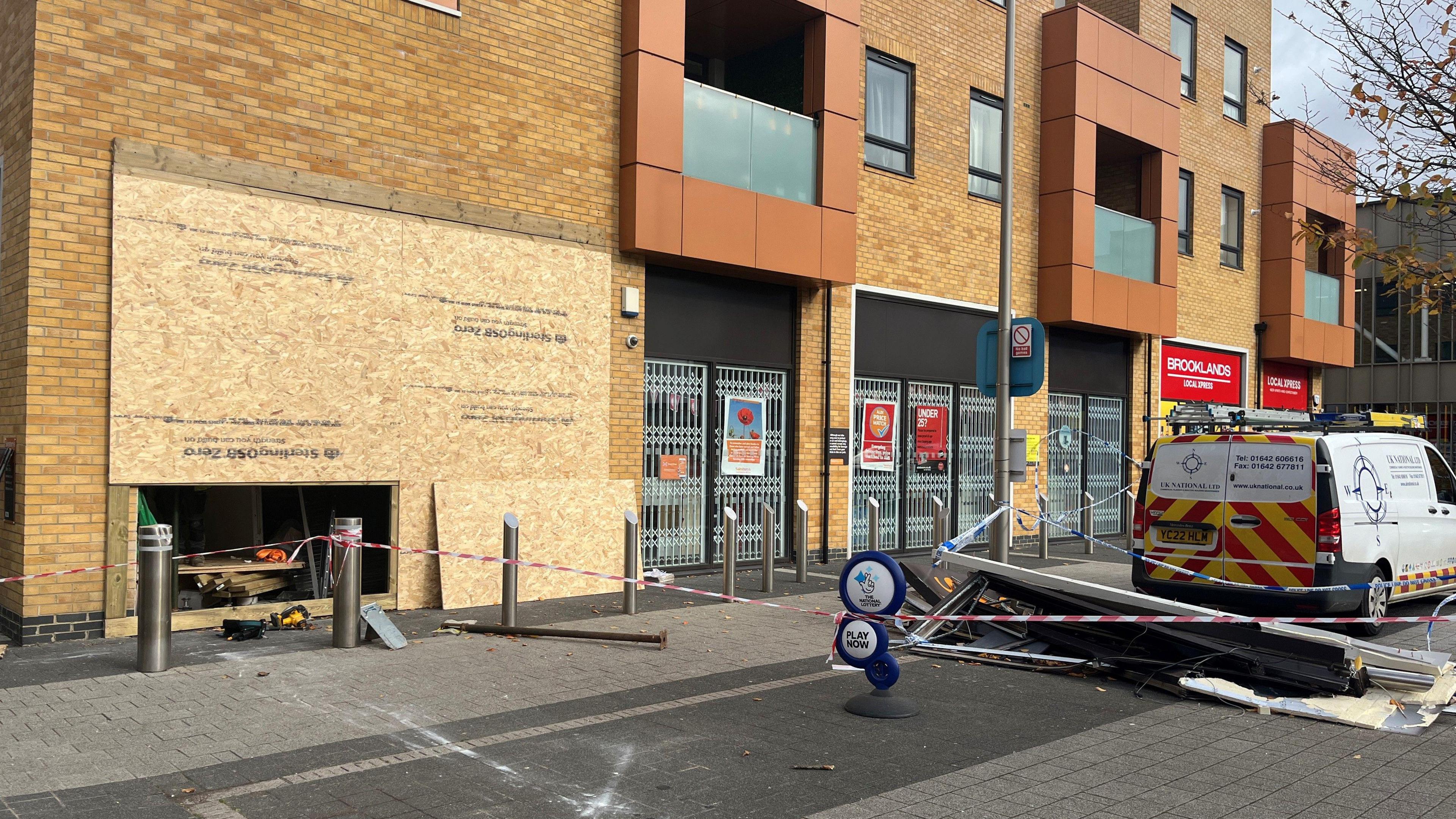 The outside of a Sainsbury's Local convenience store which has been boarded up. There is a cordon in front of the building and damaged signage of the shop laying on the ground.