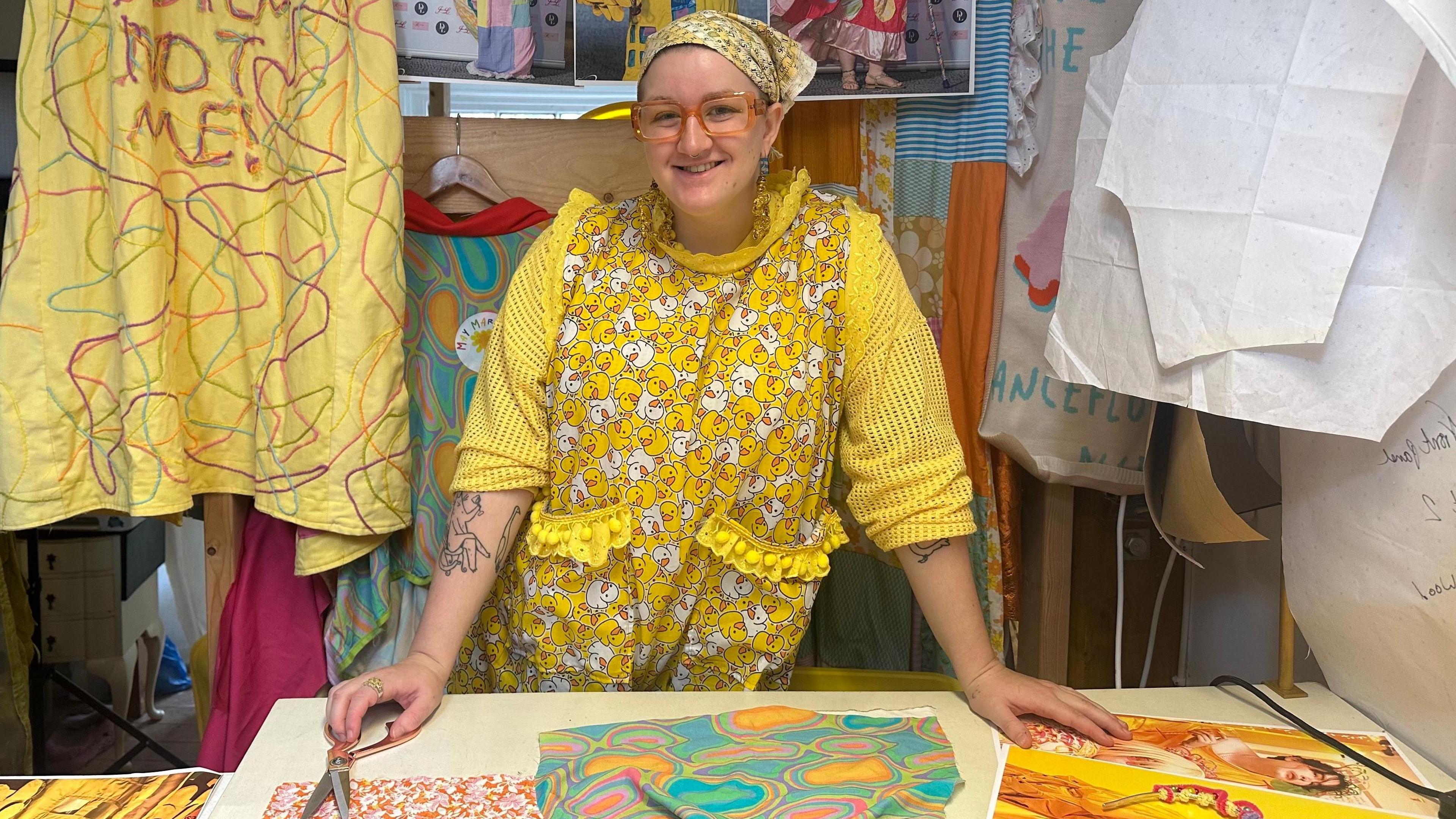 May Gauntlett leaning on her work table with a pair of fabric scissors under her right hand. She is wearing a variety of yellow, patterned clothing, including a headscarf. Fabric and design images are laid on the table and hanging behind her