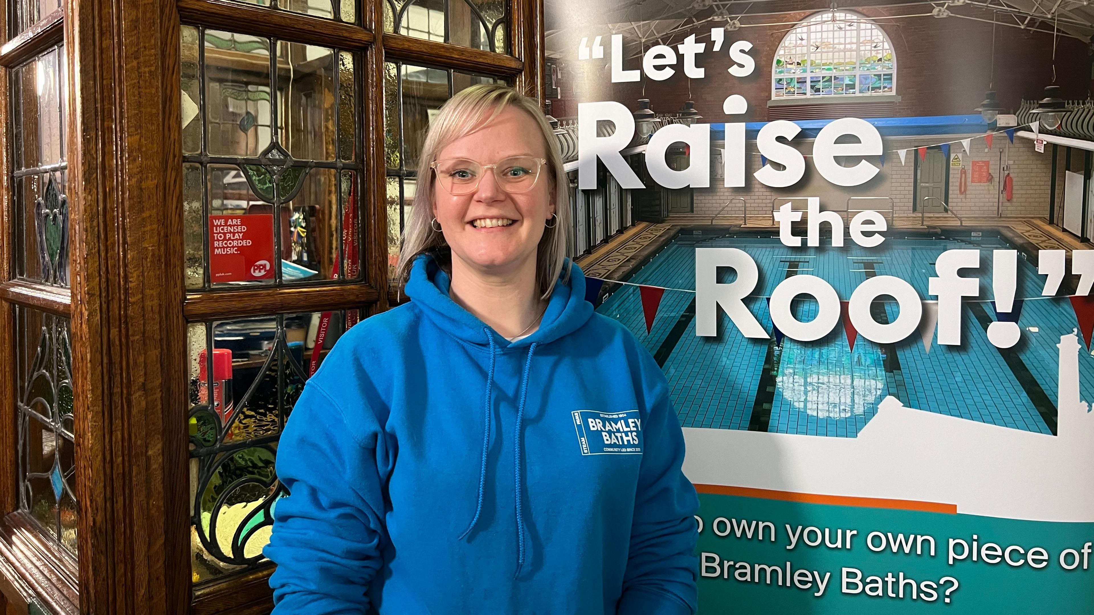 Jennie Willetts wears glasses and a blue Bramley Baths hoodie. She stands in front of a banner reading: "Let's raise the roof!".