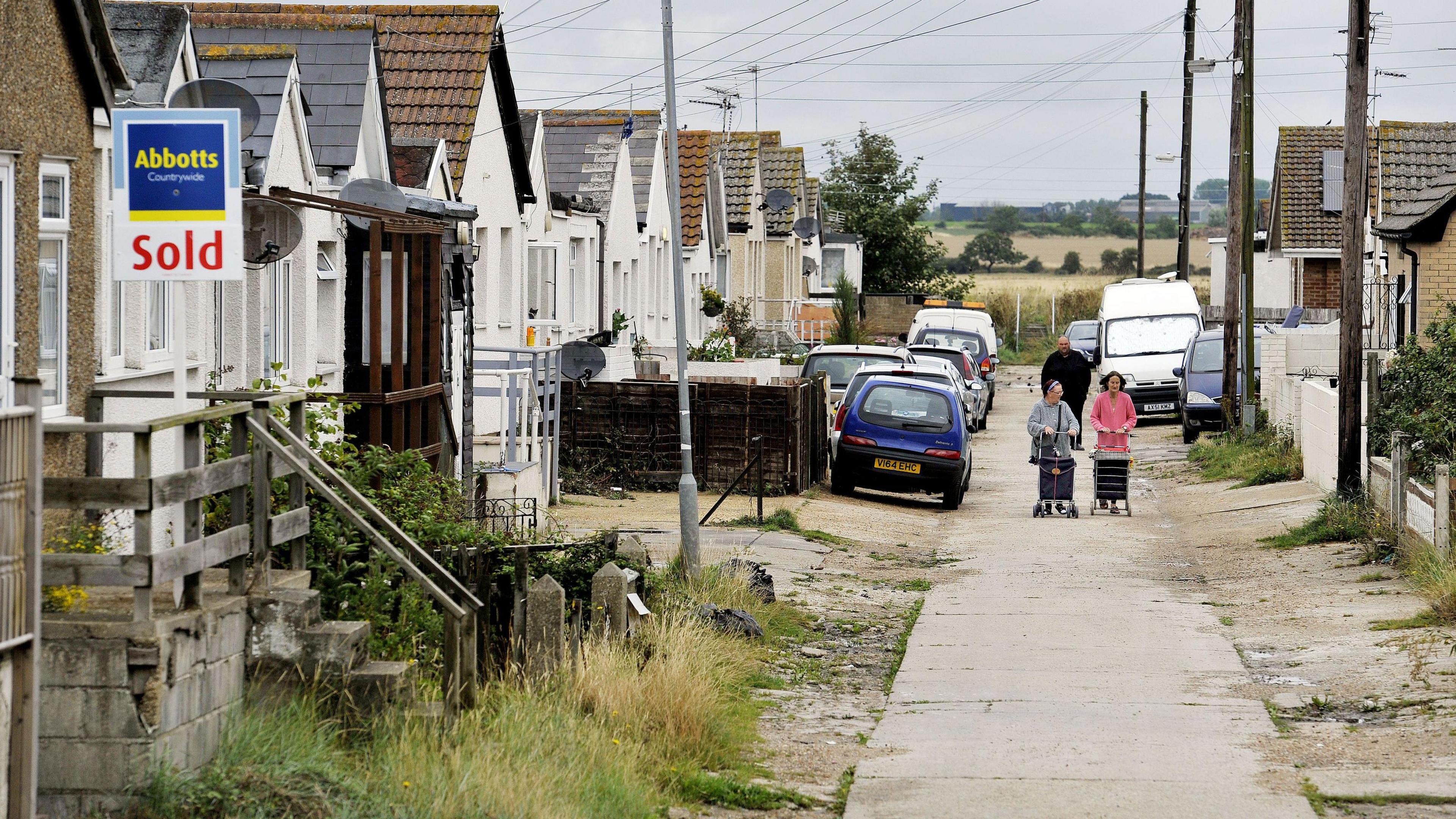 A roadway on the Brooklands estate in East Jaywick. Chalet or bungalow style homes are in a row, and two women wheeling trolleys are walking towards the camera. There is a field in the background.