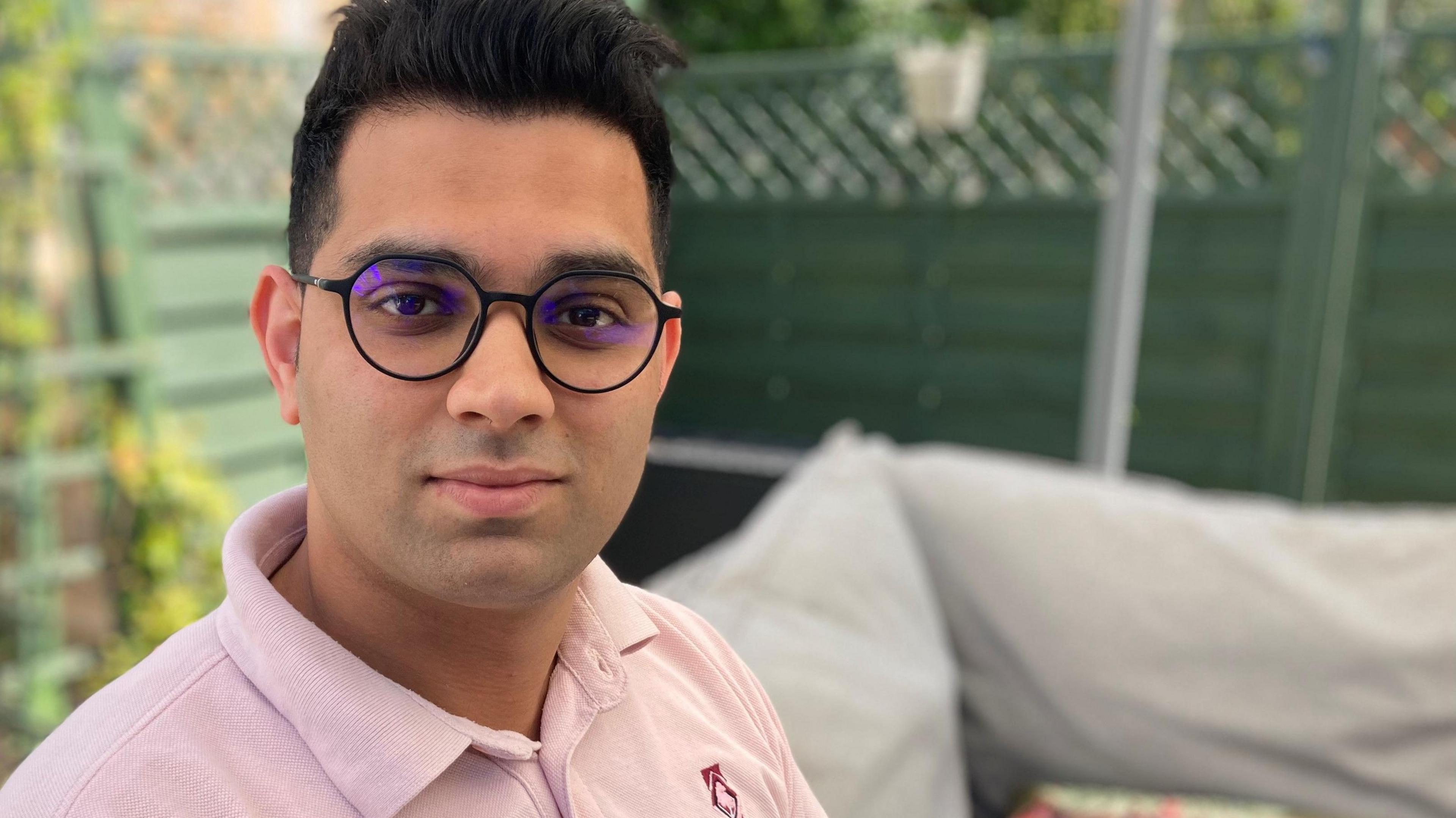 Man with short hair, glasses and pink polo shirt. He is sat on a light grey sofa. There is a patterned cushion and a green fence with plants and gravel in the background.