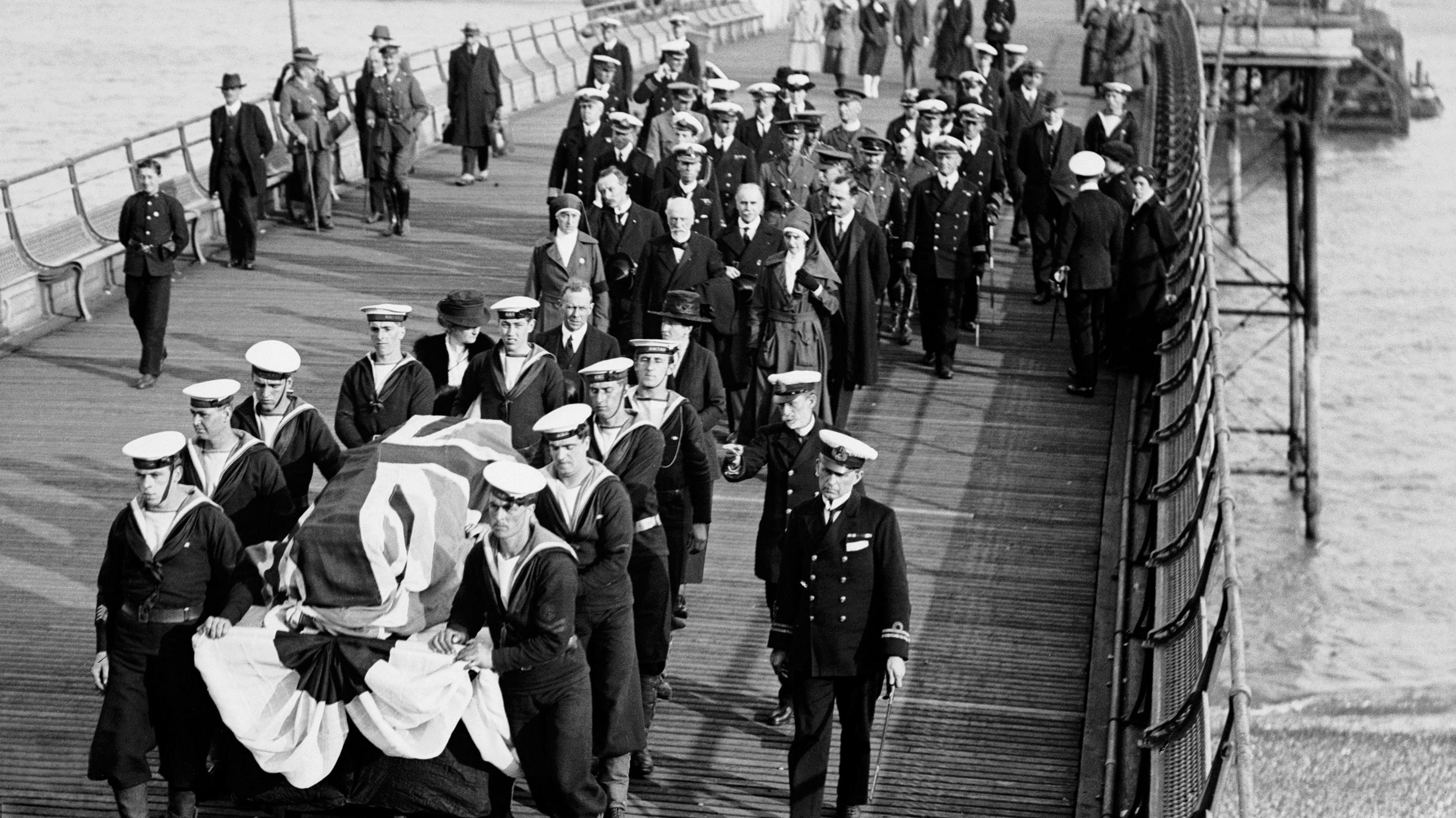 A coffin covered in a union flag is carried on a gun carriage up a pier by several sailors, with a large group of nuns and officers following behind.