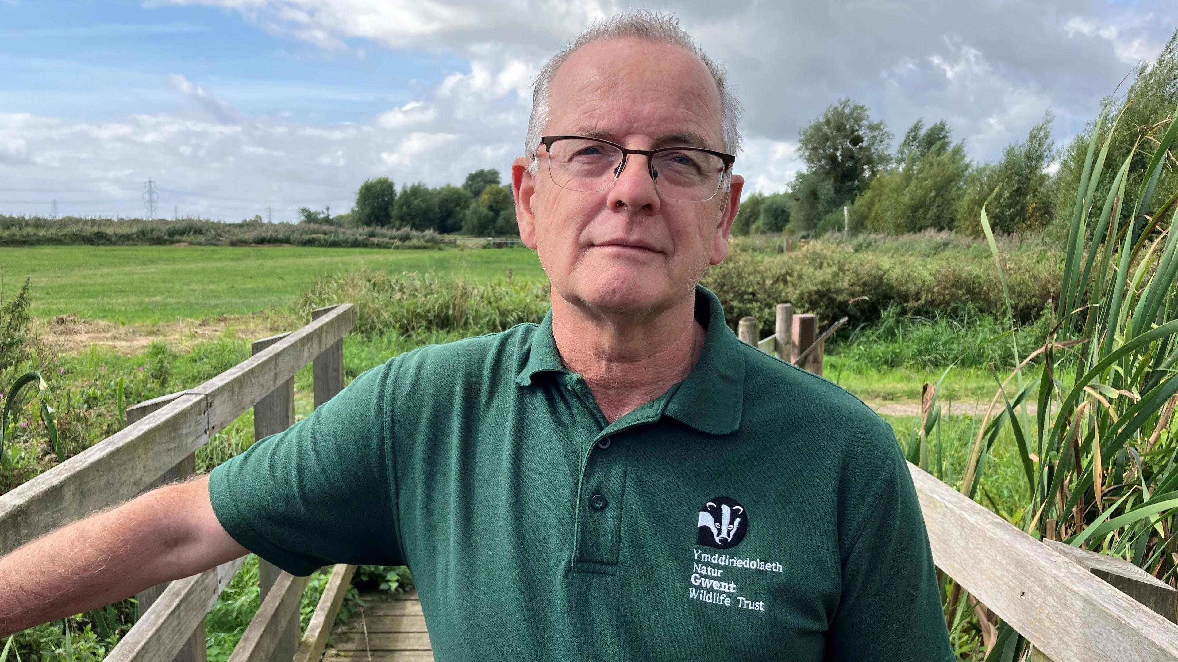 Mike Webb o Ymddiriedolaeth Natur Gwent yn sefyll ar bompren yn gwisgo crys polo gwyrdd tywyll gyda logo Ymddiriedolaeth Natur Gwent arno.