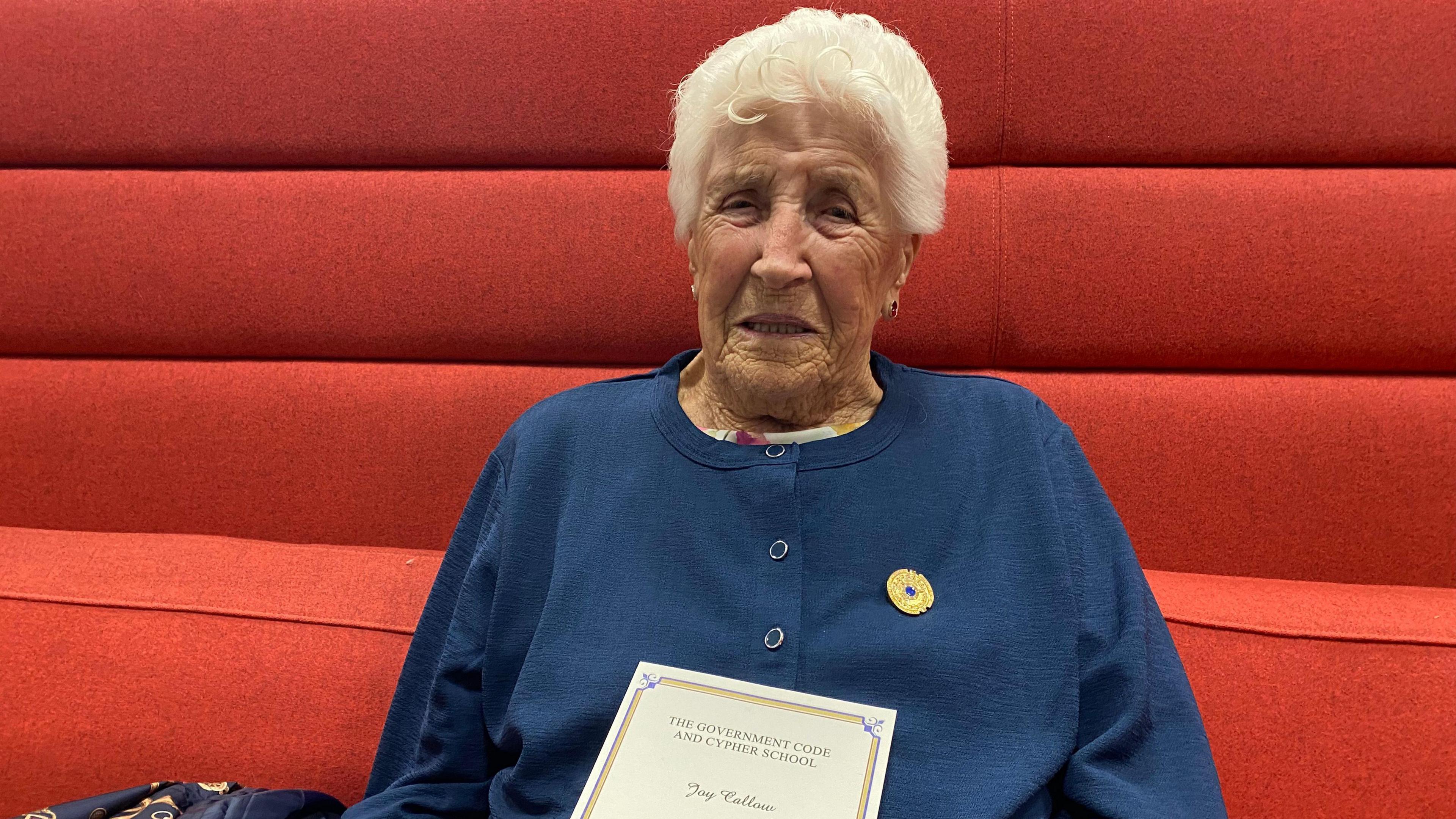 Joy Trew smiles at the camera wearing her gold and blue Bletchley medal and holding a note from the Prime Minister's office