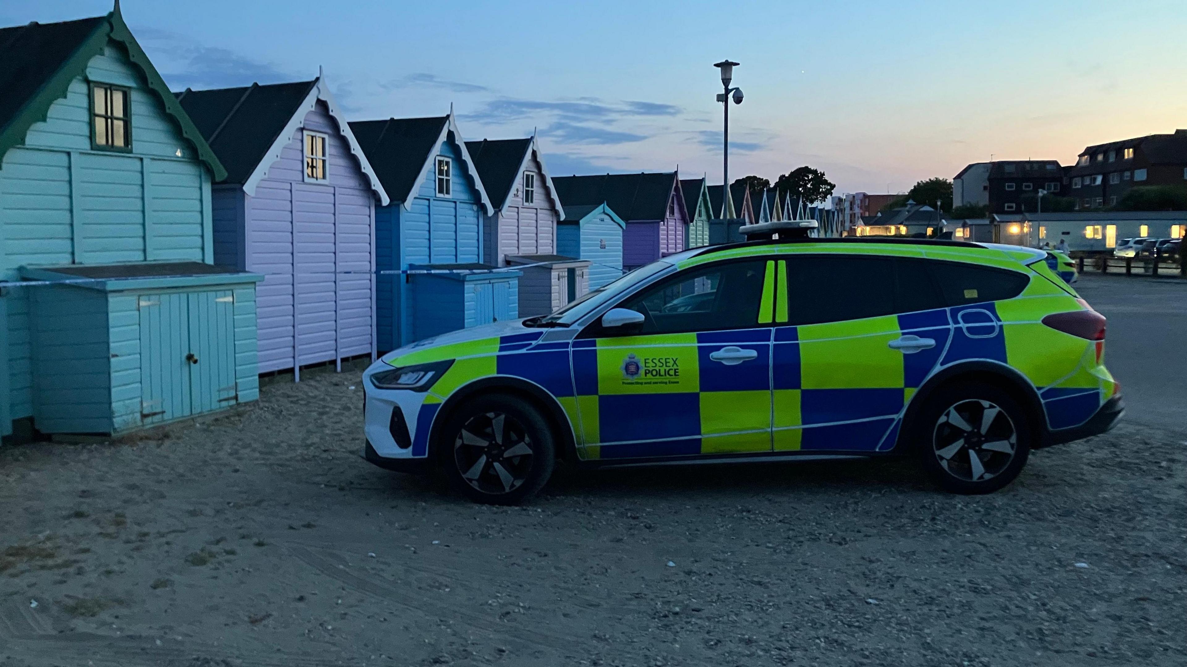 An Essex Police estate car parked on a beach. There are colourful beach huts to its left and behind it are buildings, whose lights are on. 