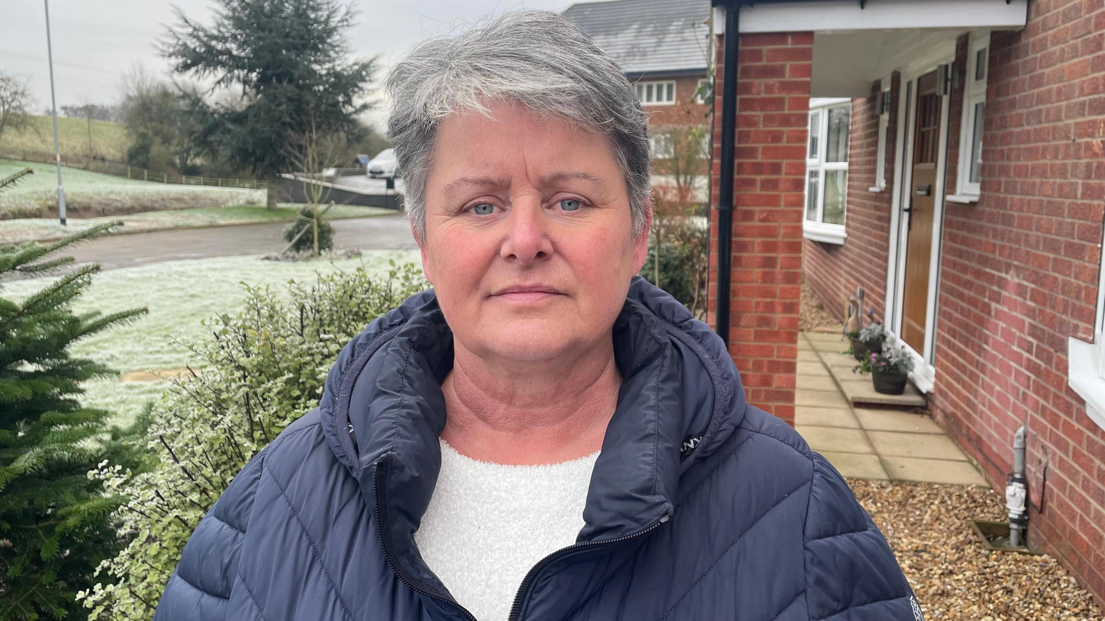 A woman with short grey hair is standing outside her property and has a fed-up look about her person. She is wearing a blue coat. The ground is frosty.