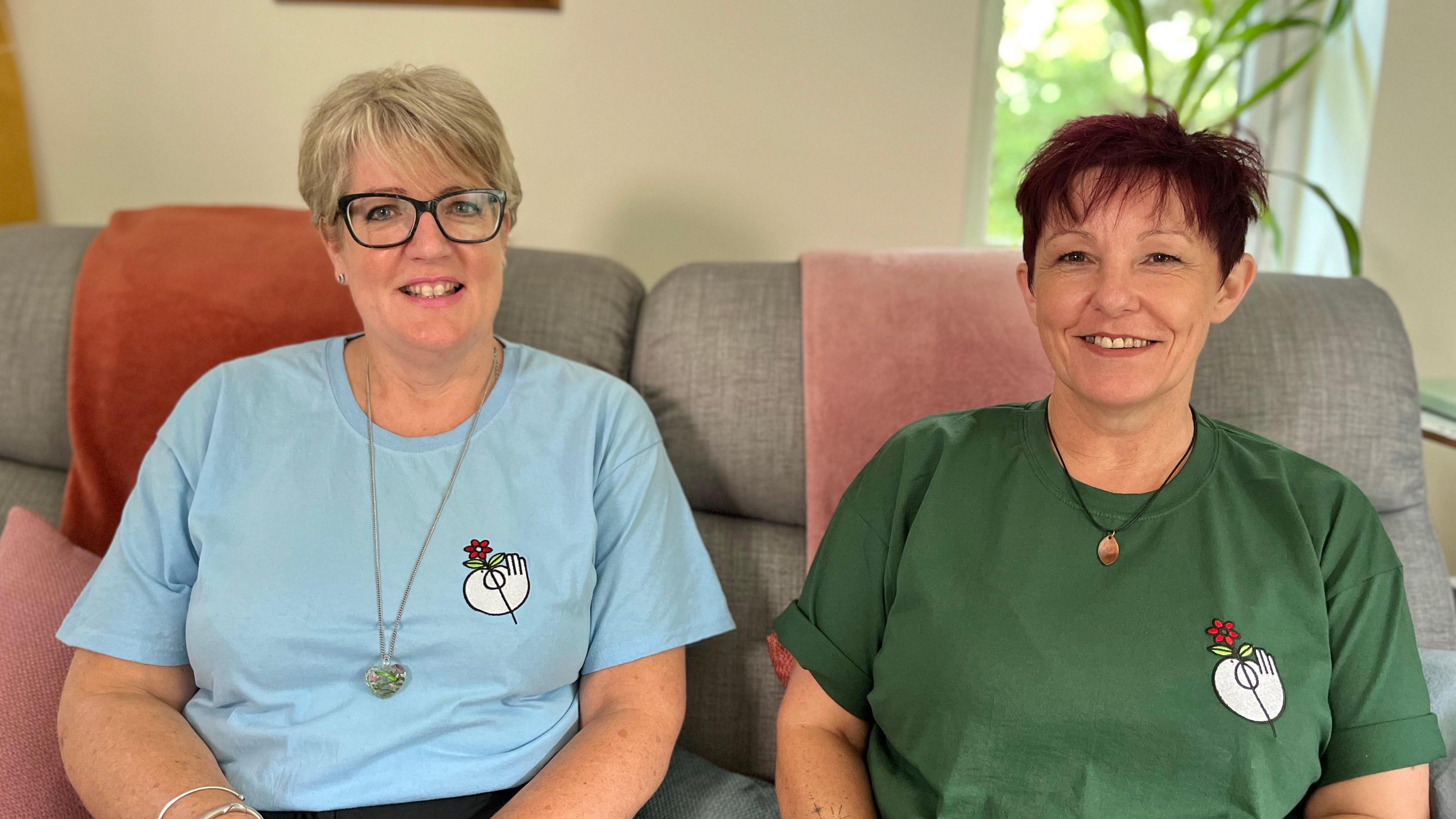 Two women sitting side-by-side on a sofa, smiling at the camera. One has short blonde hair and is wearing glasses and a light blue T-shirt. The other has dark reddish hair and is wearing a dark green T-shirt. 