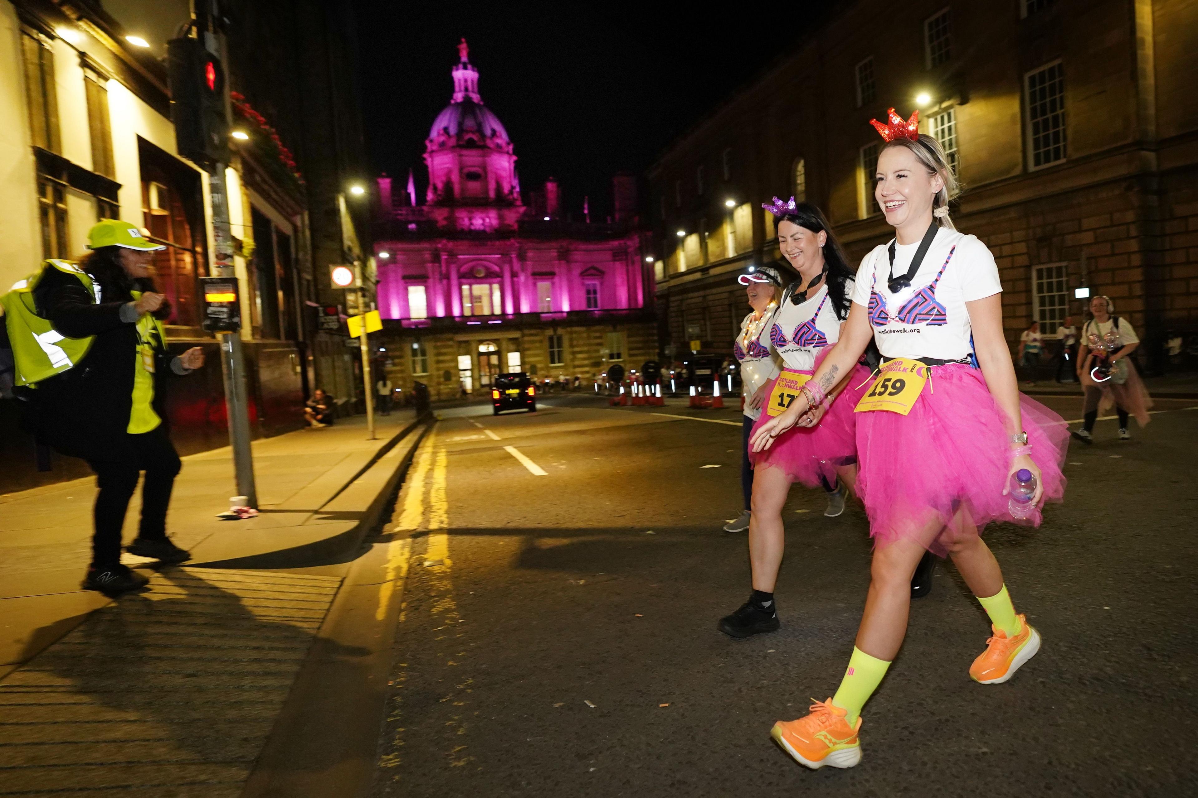 Three MoonWalkers in pink skirts and white tops cross the road in front of a building lit up in pink