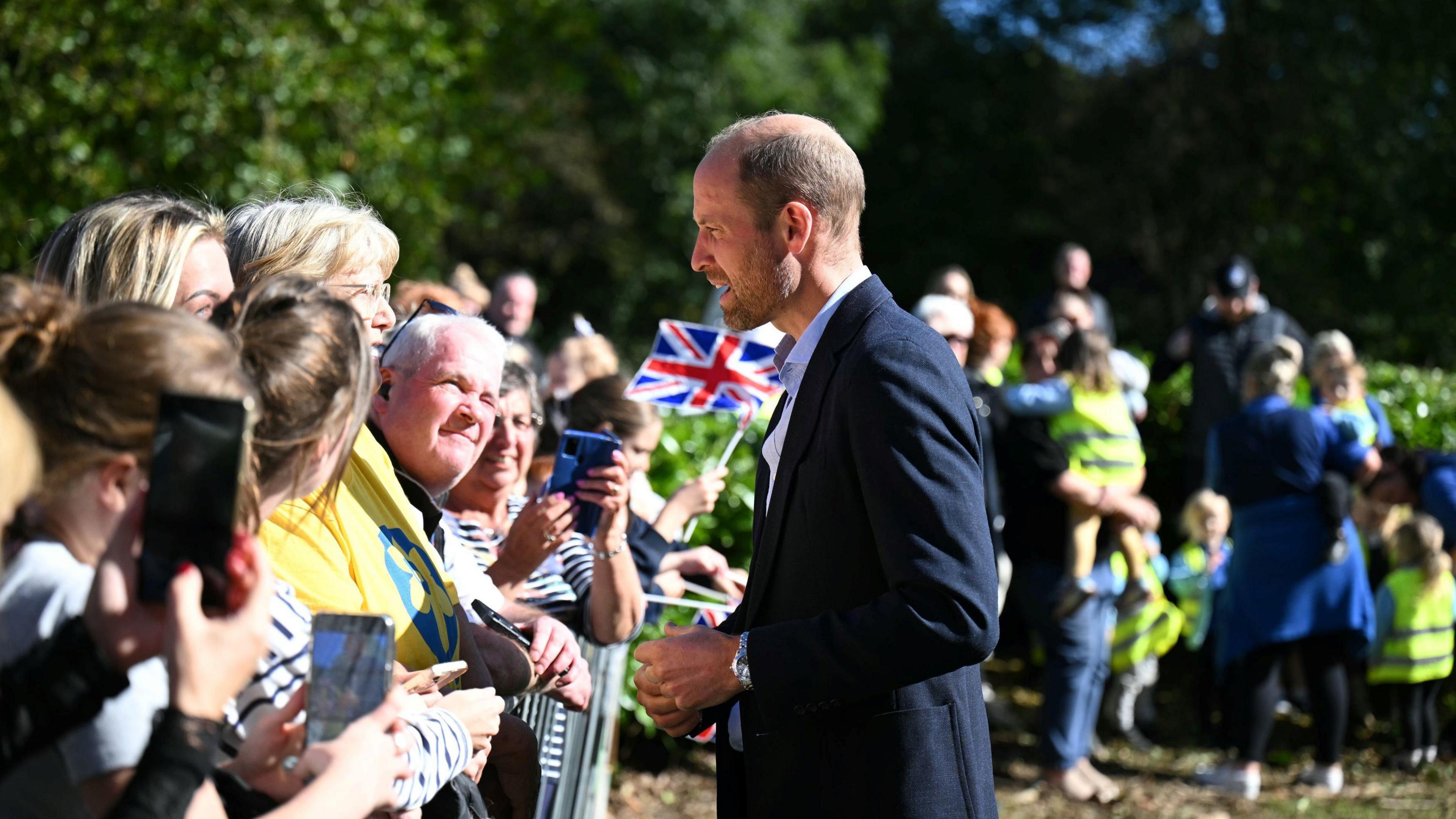 The Prince of Wales speaking with members of the public upon his arrival to visit Birtley Community Pool who are lined up behind a barrier. One is waving a Union Flag. 