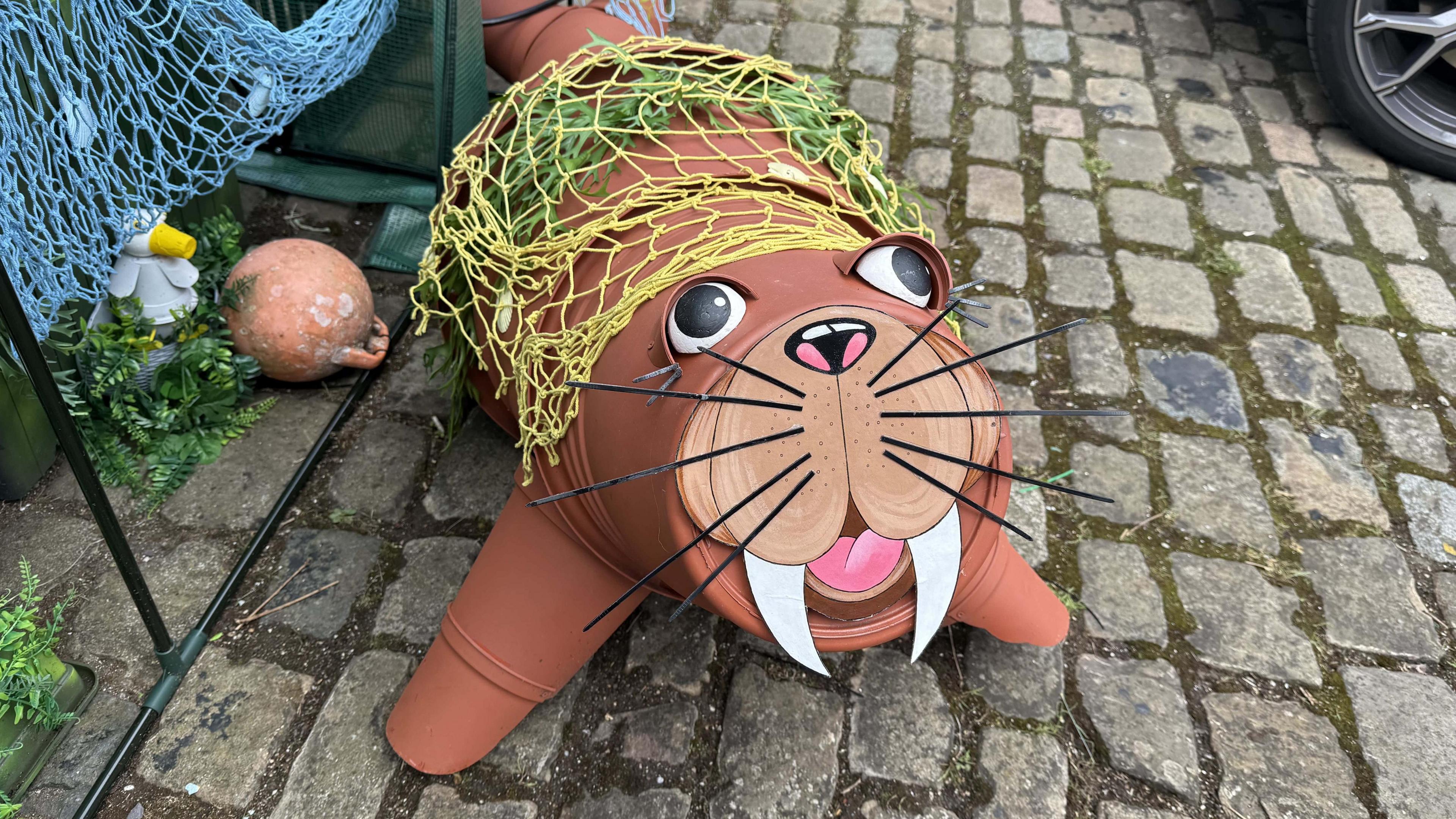A walrus with a smiling face and large tusks has been made using brown flower pots. It is laid on a cobbled path and surrounded by fishing nets.