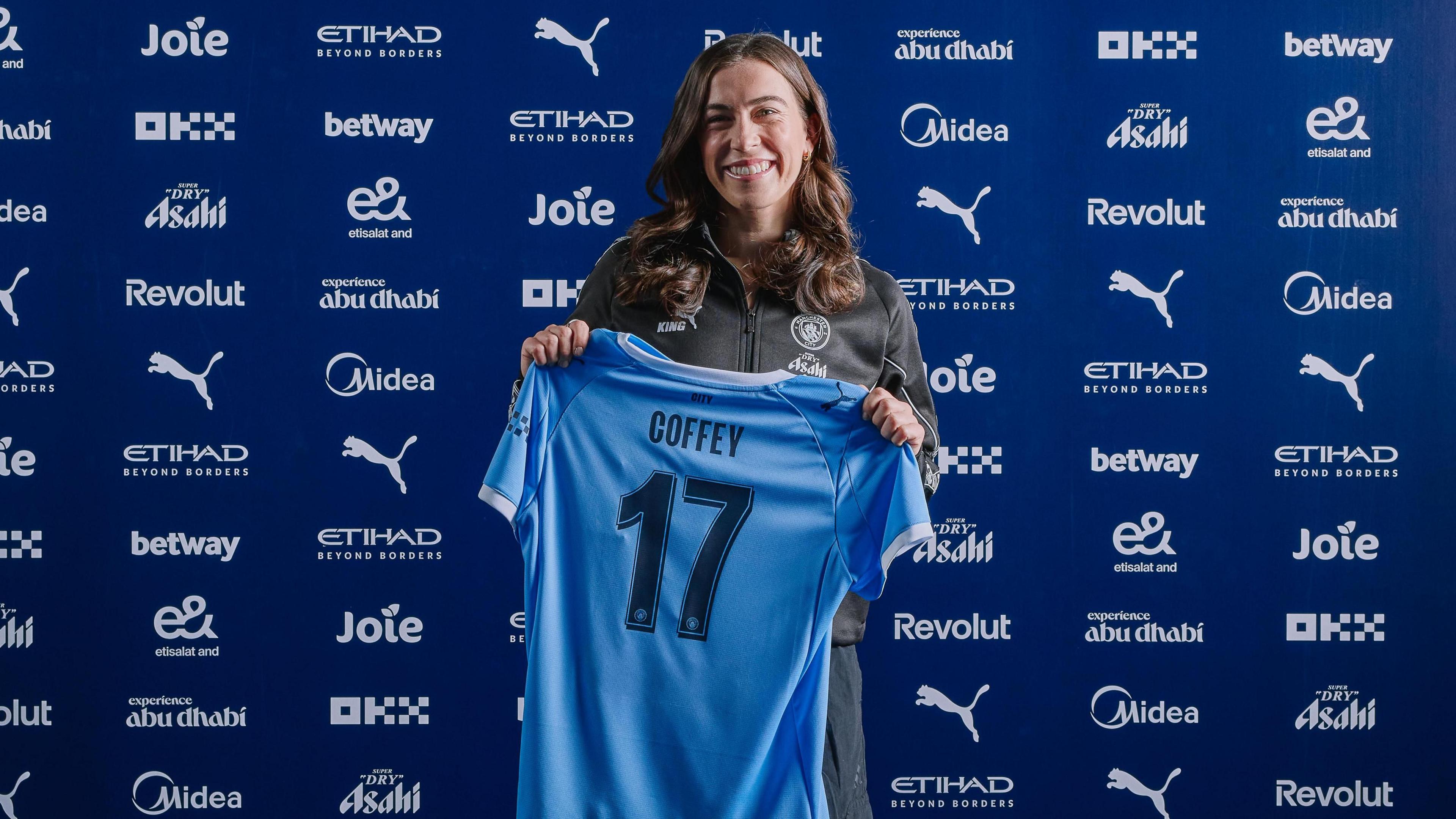 A smiling Sam Coffey holds a blue Manchester City shirt with Coffey 17 on its back, as she stands in front of a wall showing sponsor logos