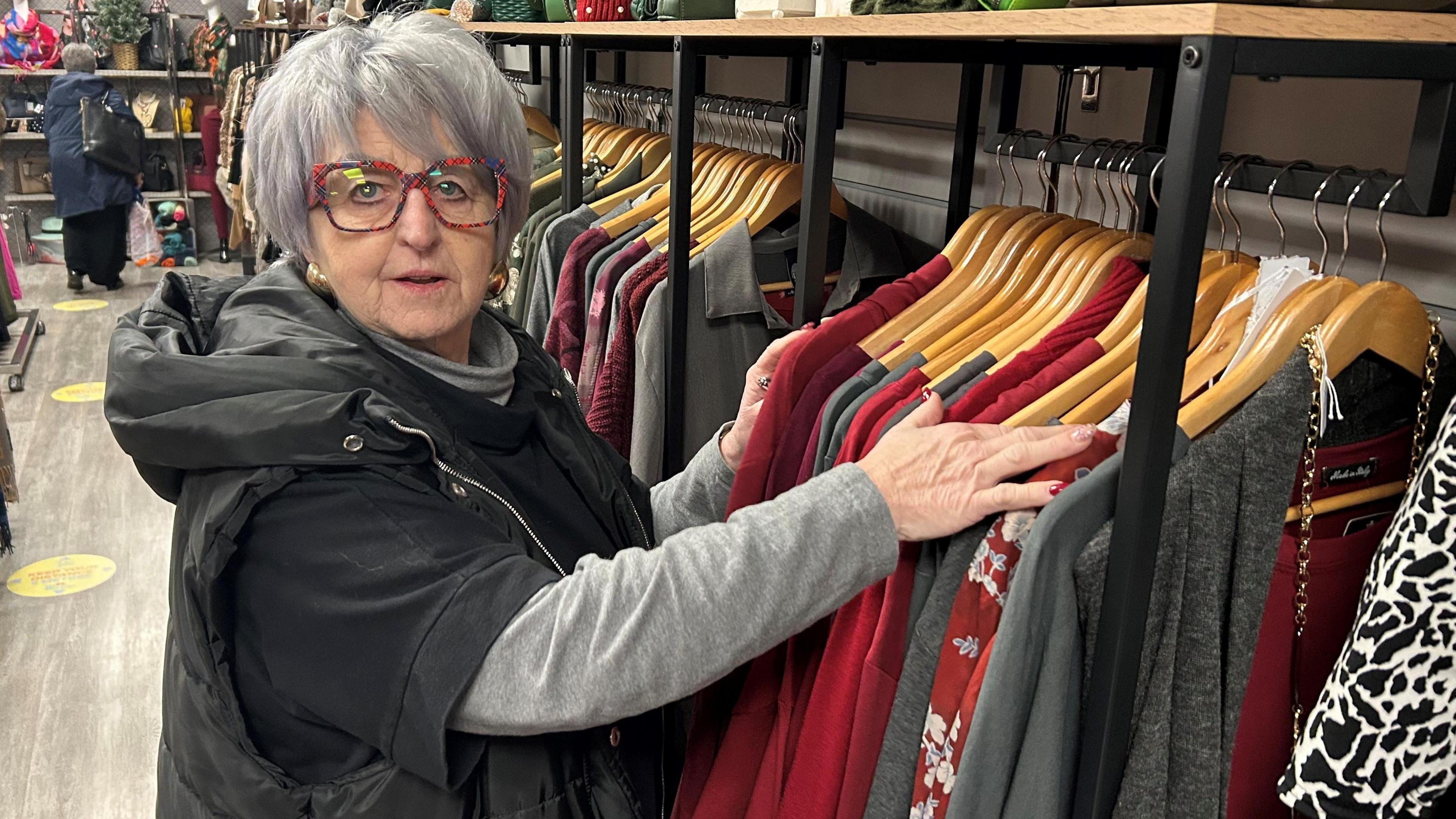 Paula Owen looking at the camera and is going through clothes on a clothes rail. She is wearing colourful glasses and has grey hair. 