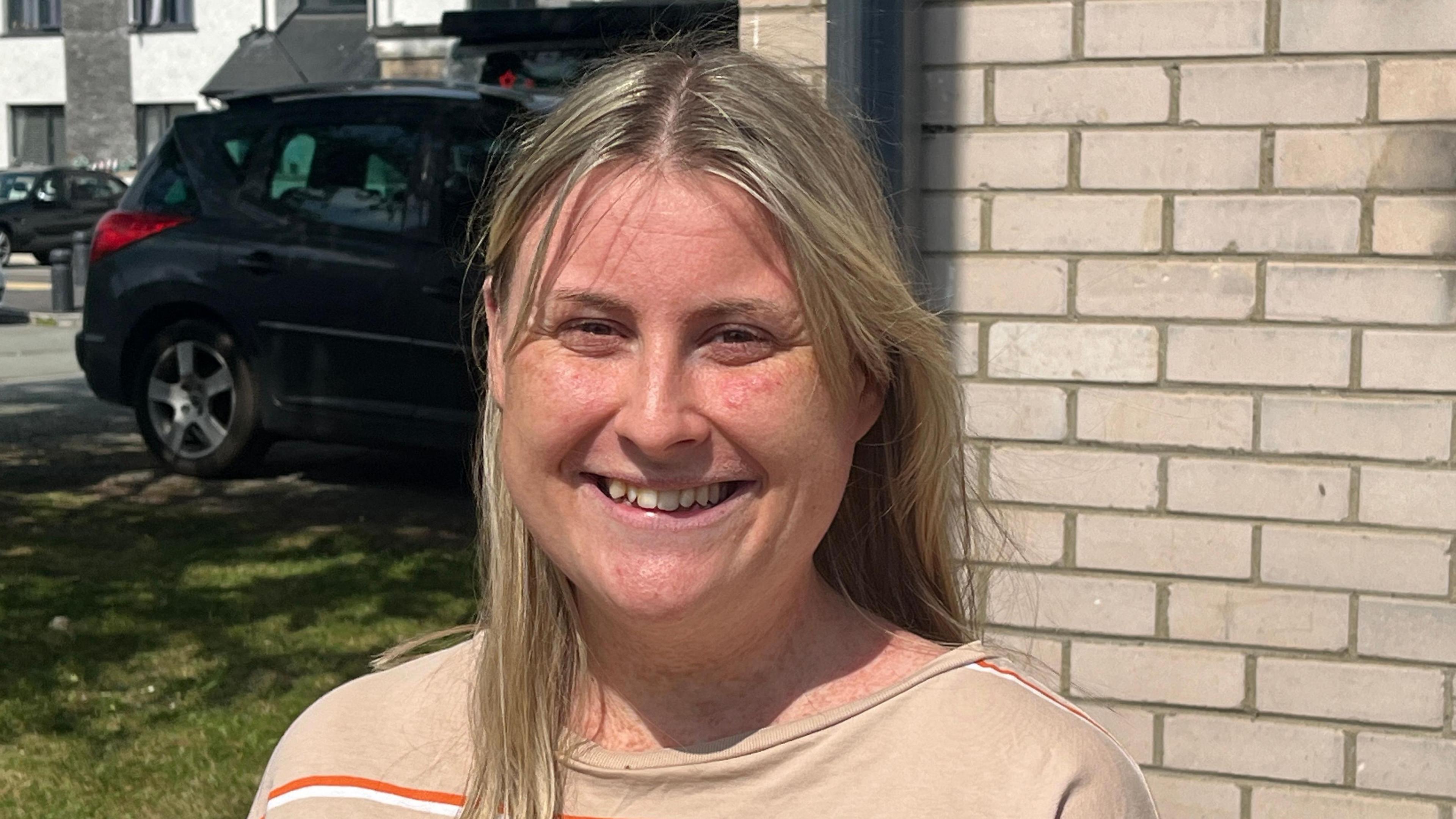 Lily Jones smiling pictured against brick house wall with lawn in the background and a black car. She has long blonde hair and wears a beige top with orange and white stripes.