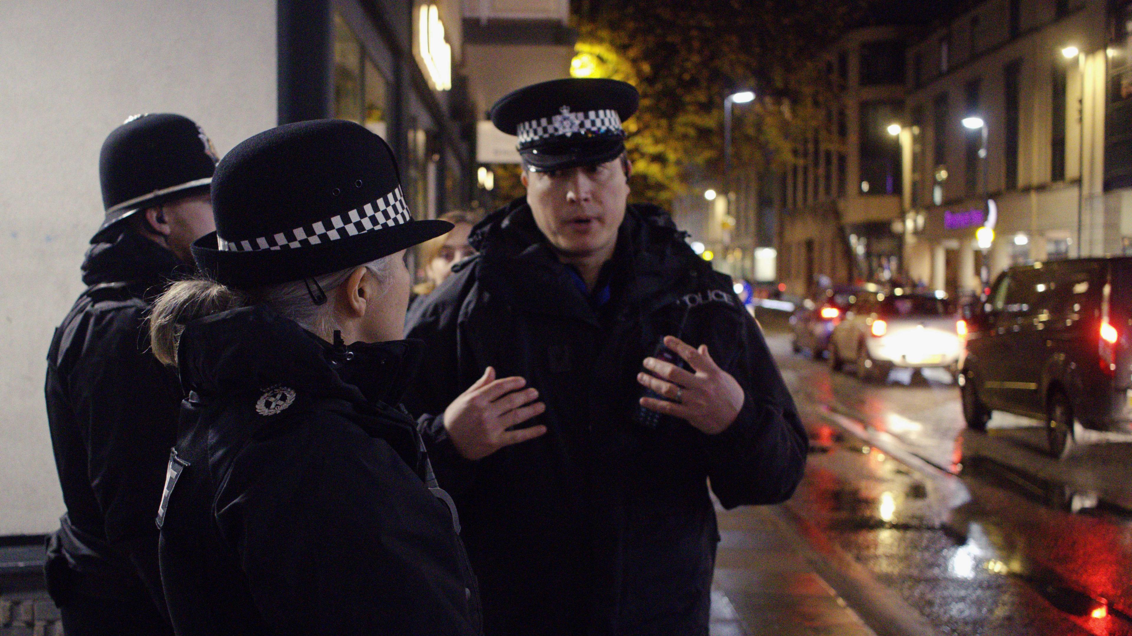 Chief Constable Crew in uniform in the foreground speaking to Neighbourhood Inspector Dudley Bond in Bath. They are on a busy street with lots of cars driving down it at night. Everyone is wearing full police uniform, including caps and hats.  