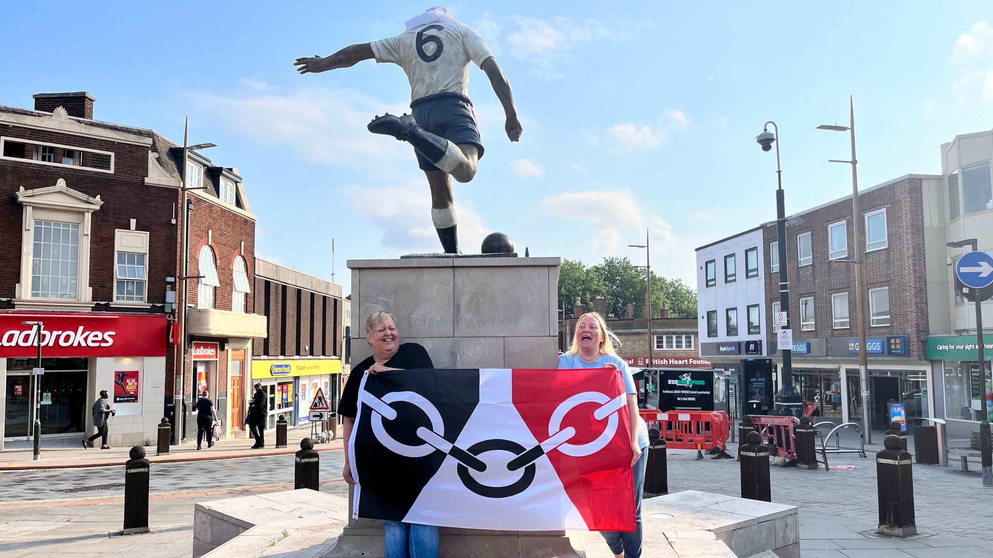Two women are standing in front of a statue in Bilston town centre. They are holding the flag of the Black Country. The flag is black, white and red with chain representing the region's manufacturing heritage.
