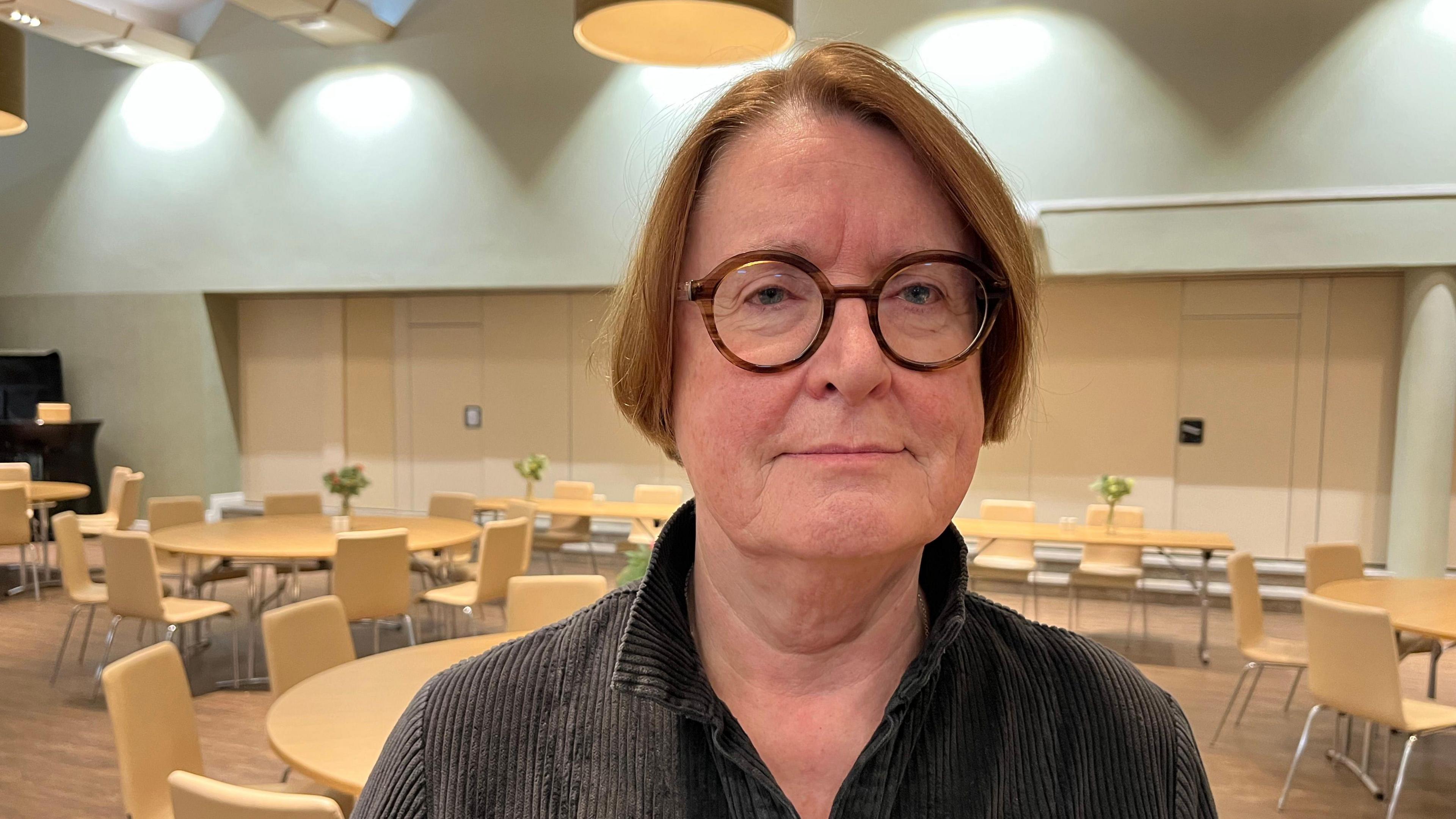 A picture of Elwen Evans looking towards the camera. She has short brown hair and brown round glasses. She is standing with tables and chairs behind her. 