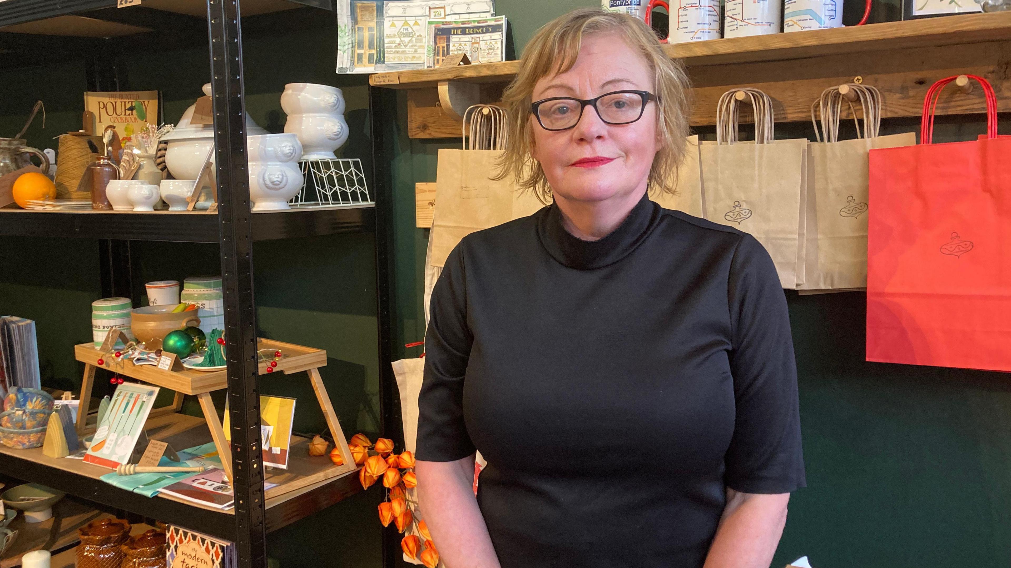 Jayne Colman standing in her shop in Pontypridd, next to shelves displaying items for sale including mugs, pots and books