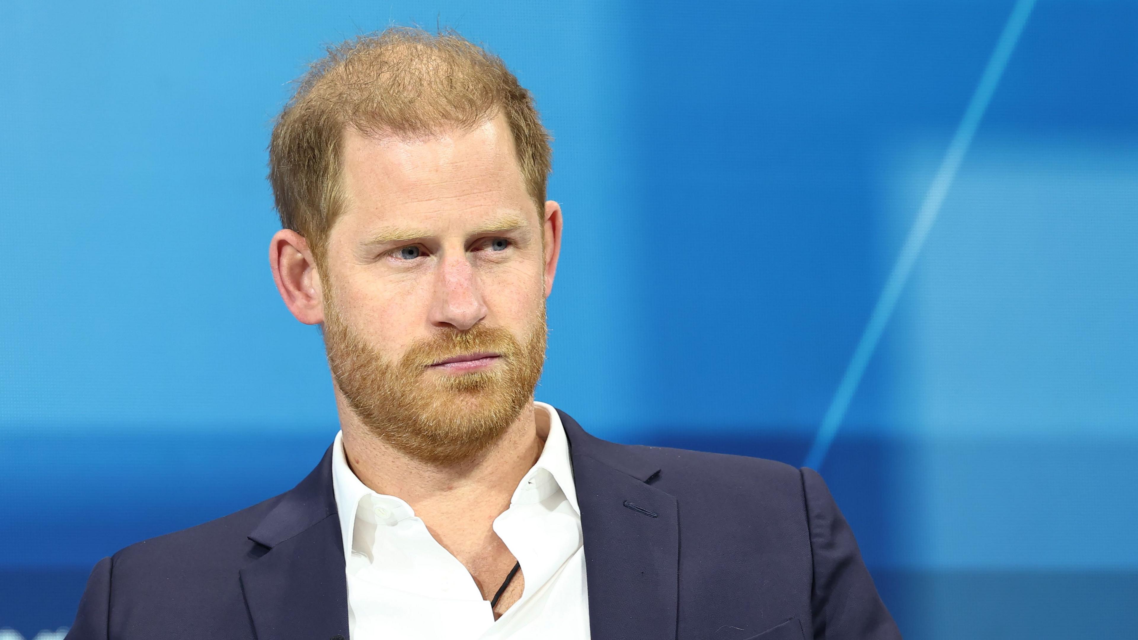 Prince Harry wearing a blue suit jacket and white shirt with no tie. There's a blue background behind him and he's looking off into the distance.