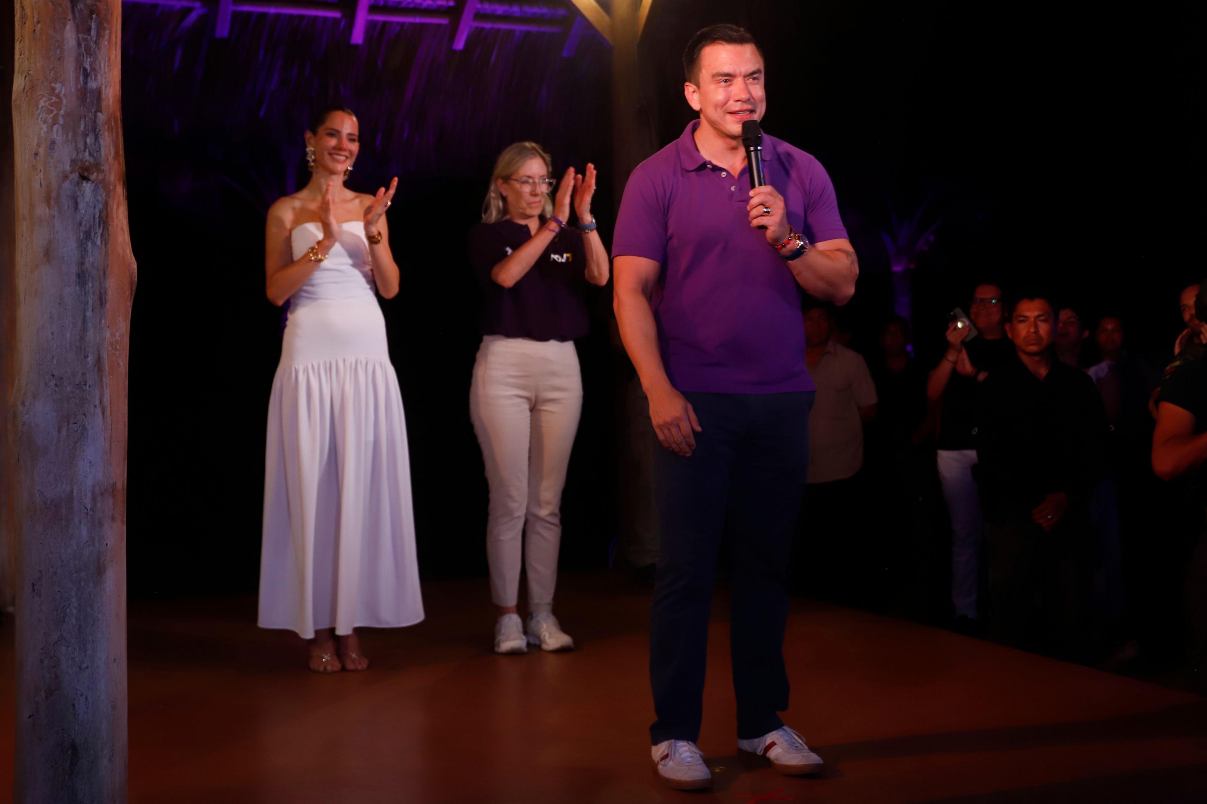 A man, Daniel Noboa, wears a purple polo shirt speaks into a microphone with two women clapping behind him