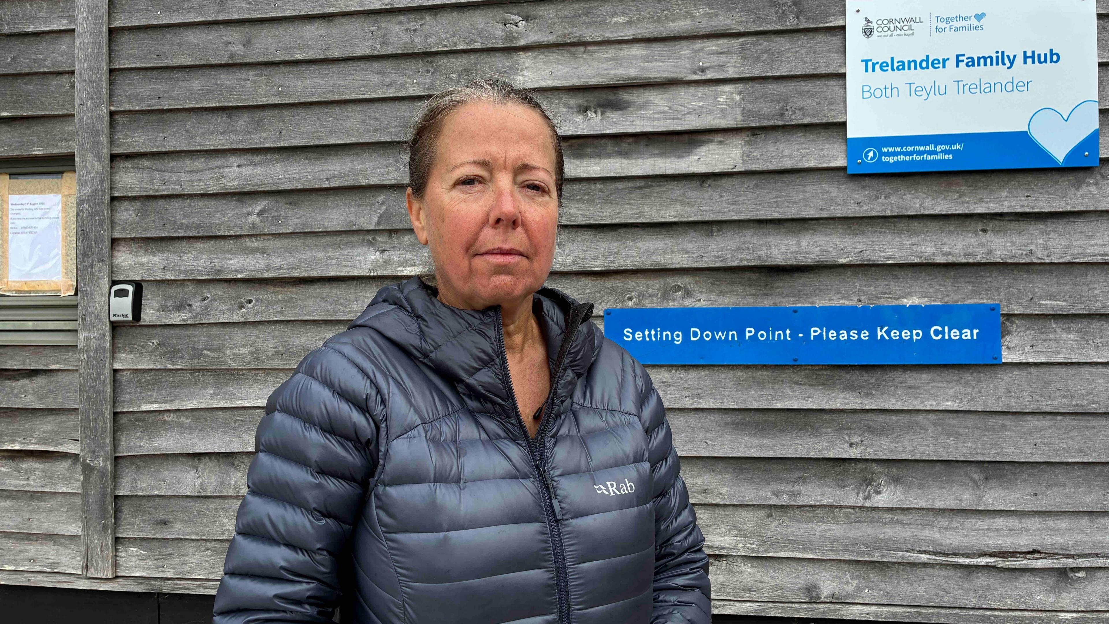 The photo shows a grey-haired woman in a puffer jacket stands in front of a slatted wooden wall of a hut.