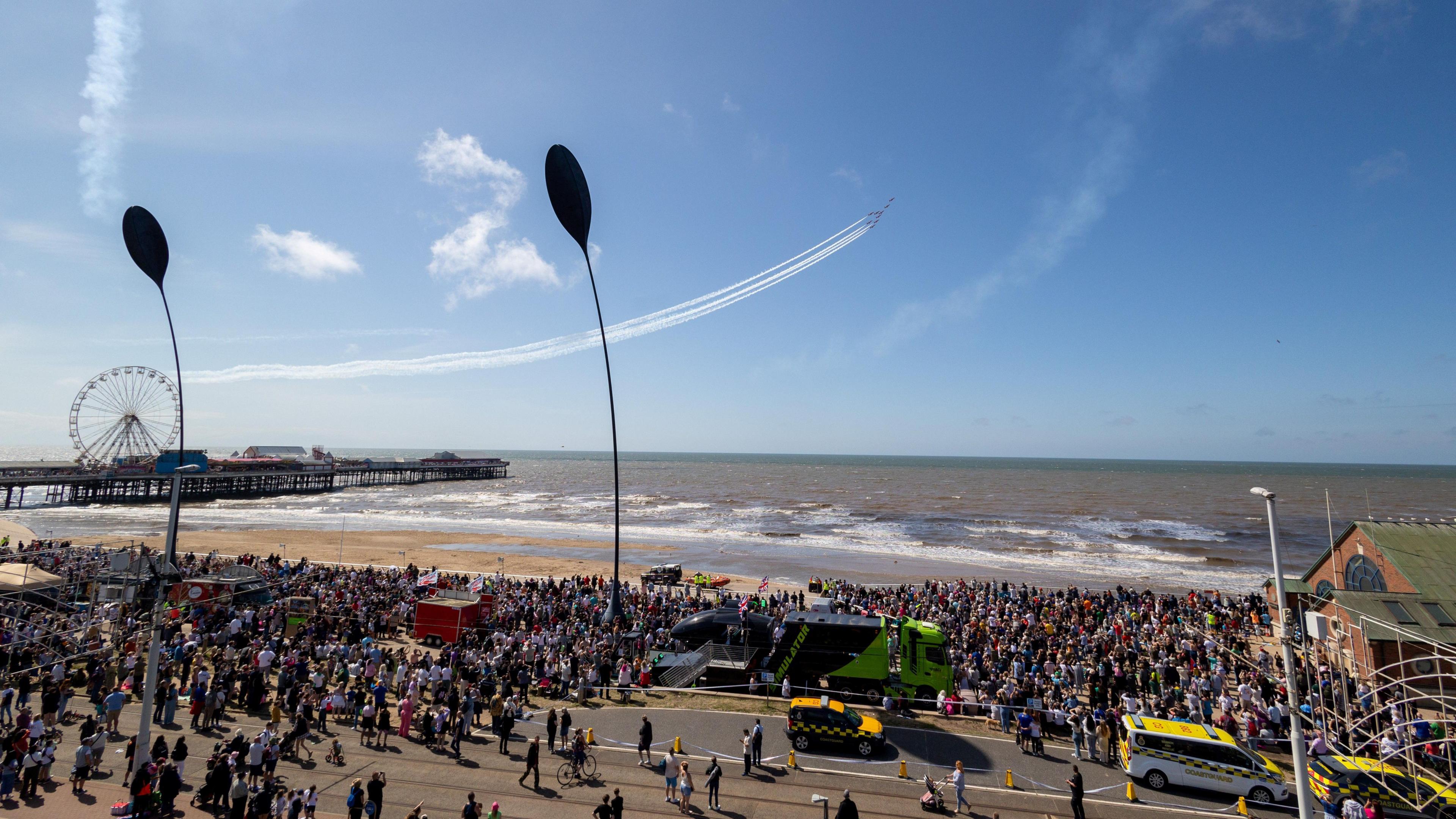 a wide shot of blackpool beach, with thousands of spectators. on the right of the beach there is a building made of red bricks and grey roof. Above the sea, in the middle of the picture there are some planes flying, trailing white vapor behind them. On the left, there is a pier stretching out to sea with a ferris wheel.