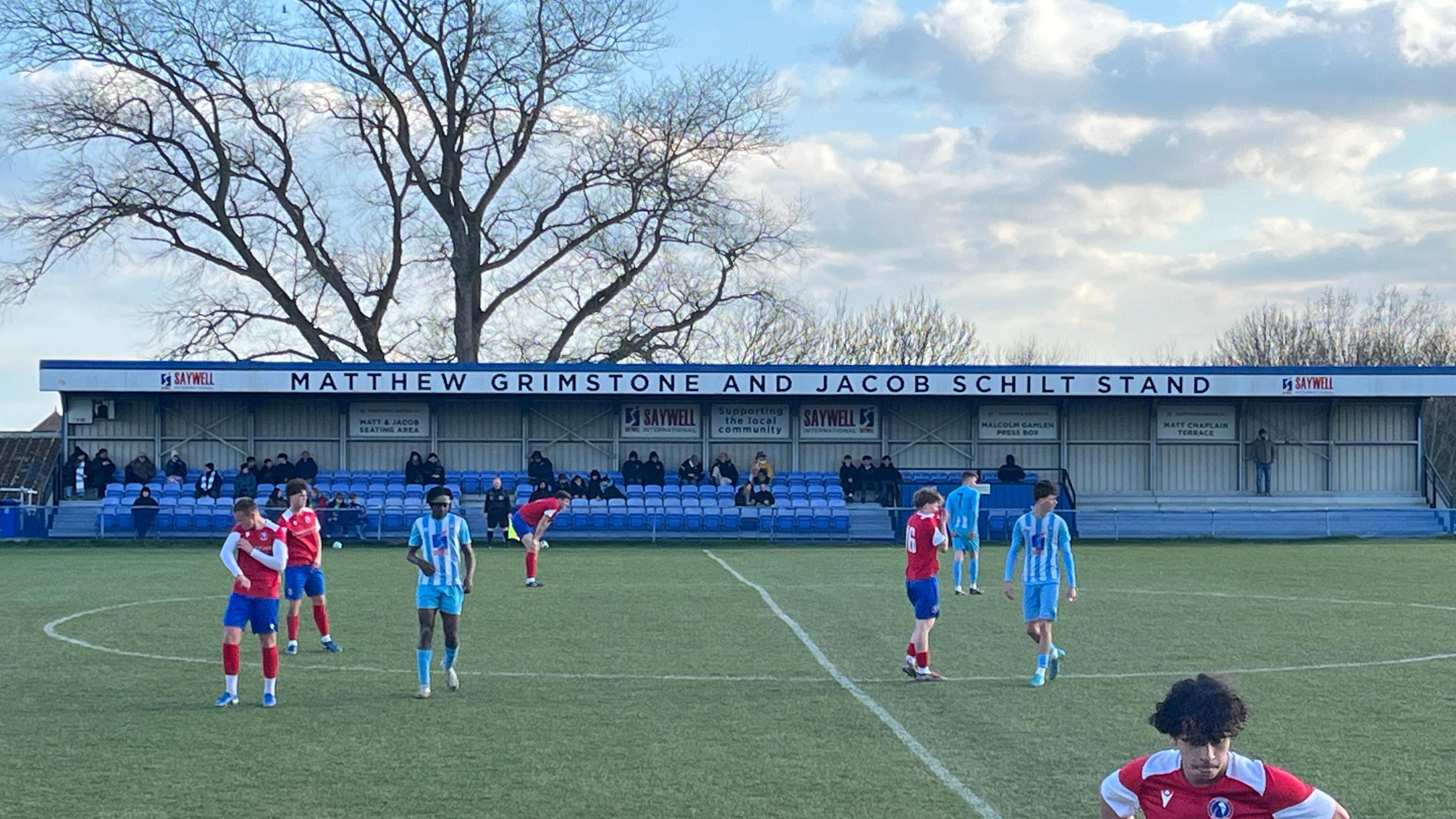 A football stand has the words "Matthew Grimstone and Jacob Schilt stand" at the top. There are people seated, watching a match, and footballers play in front of them. A large tree rises above the stand. the sky is blue with some white clouds.