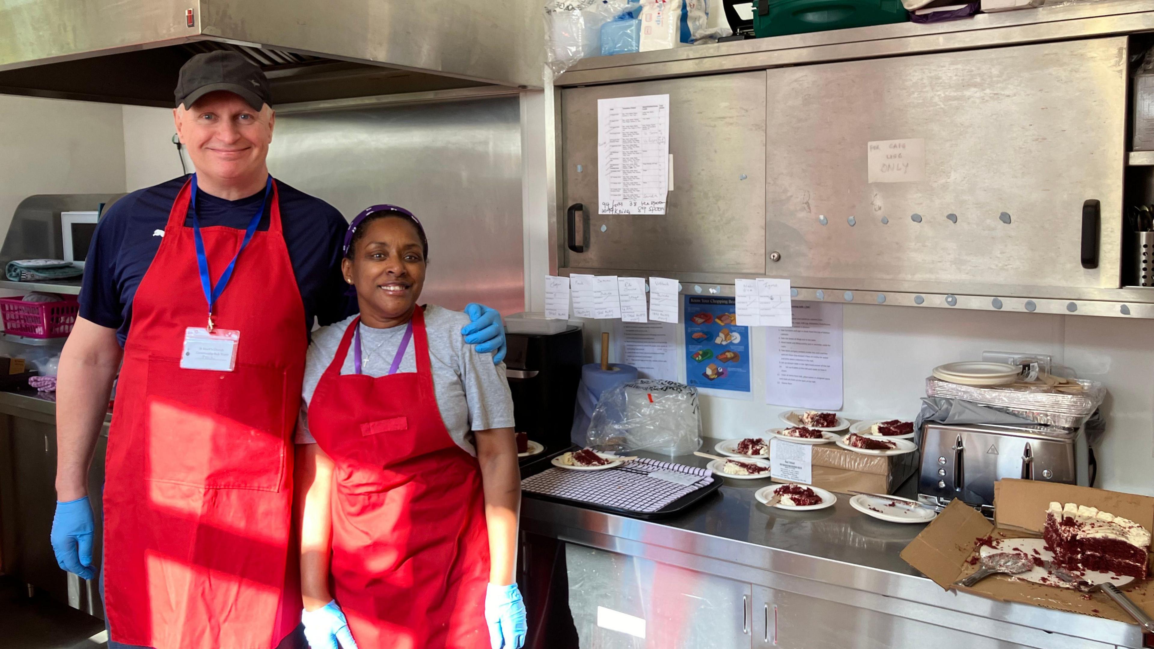 Kitchen volunteers wearing a red apron standing next to a worktop smiling for the camera 