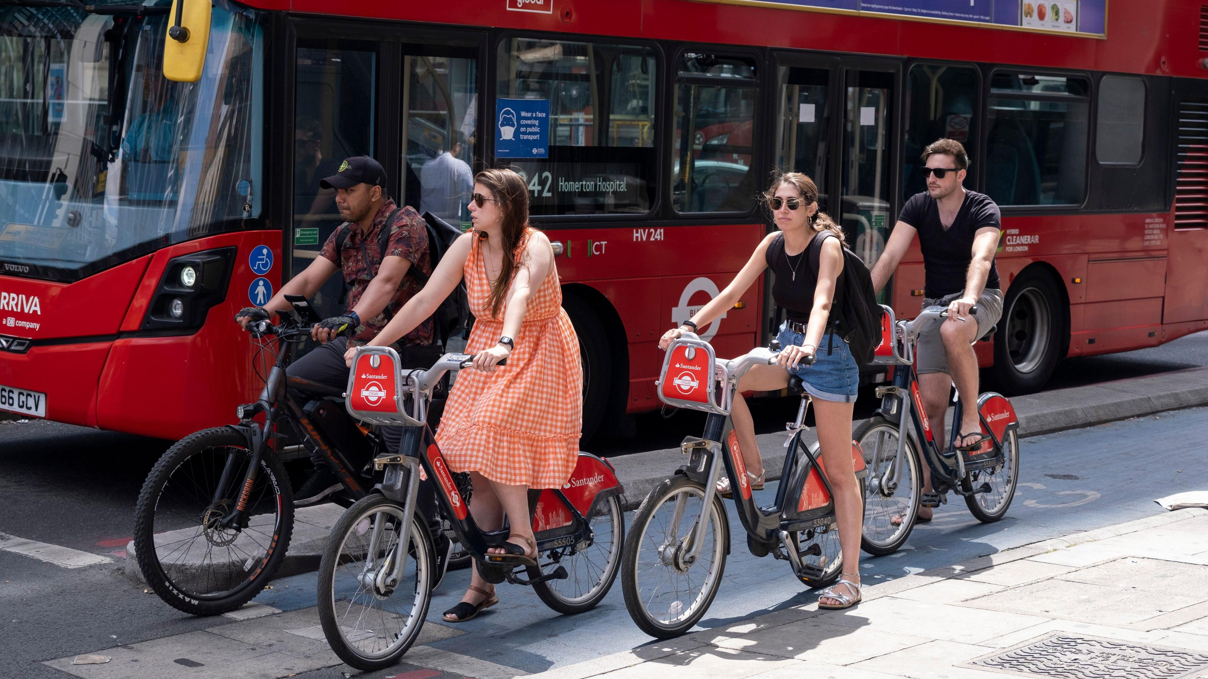 Group of four cyclists using red Santander bikes stopping at the traffic lights. Alongside them is a red London bus.