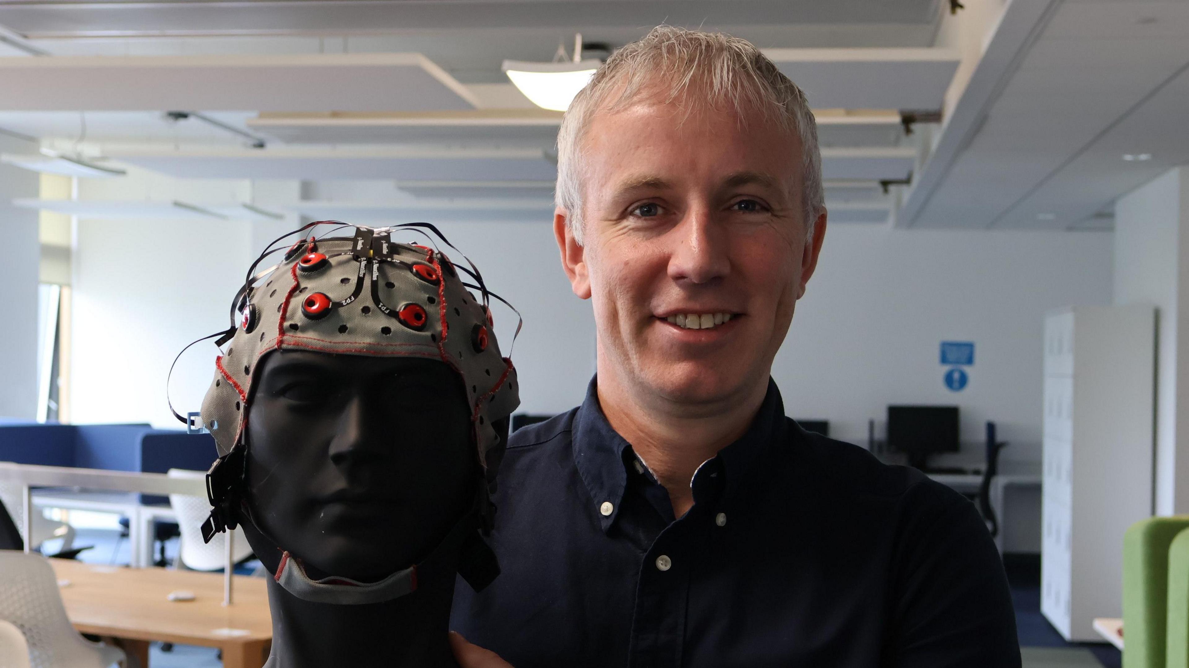 Professor Damian Coyle wearing blue shirt and holding a headset on manikin standing in university classroom.