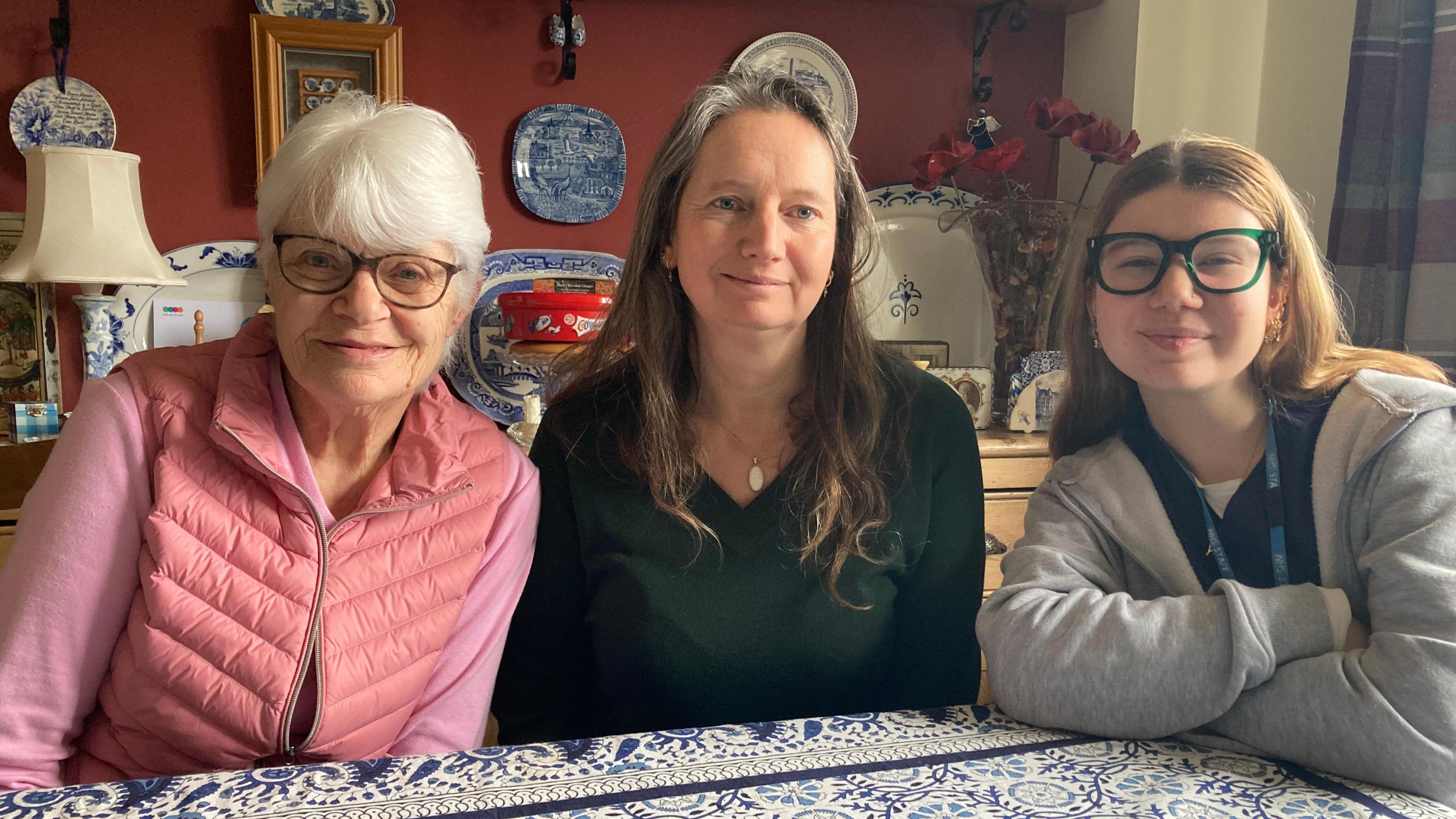 Three women sit at a kitchen table looking at the camera. One is wearing a pink gilet, the middle one wears a green jumper and the third, a teenager, wears a grey sweatshirt and green glasses. The kitchen is decorated with blue and white china and a blue and white table cloth. 