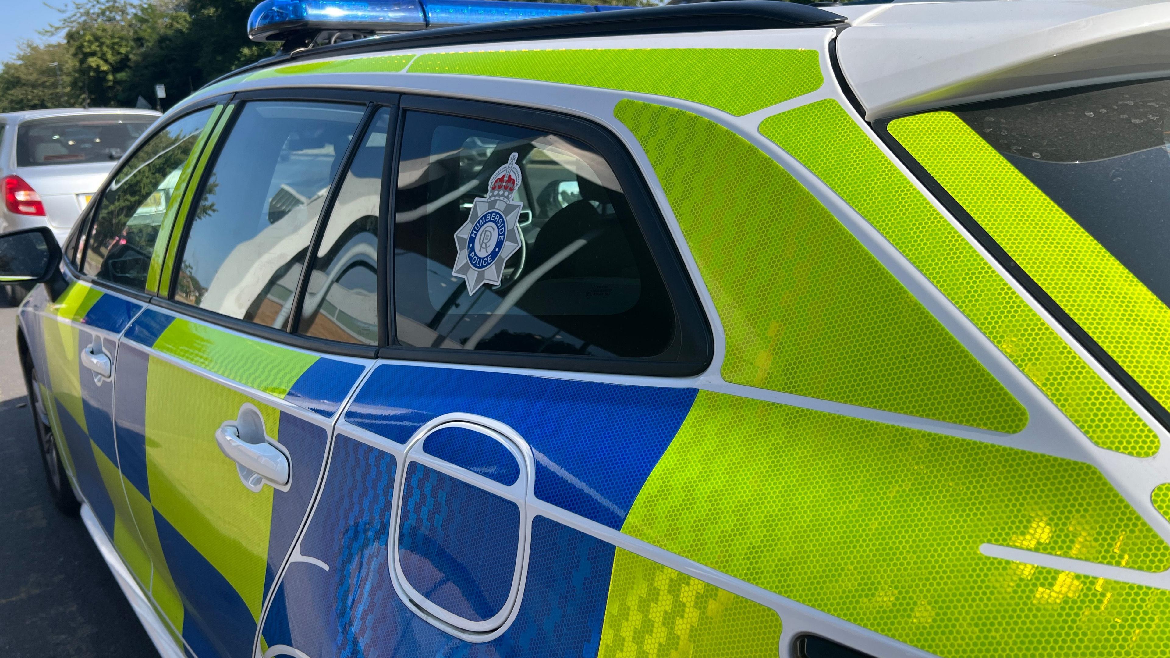 A close-up of the passenger side of a police car marked with blue and luminous yellow livery and a Humberside Police insignia in the rear side window.