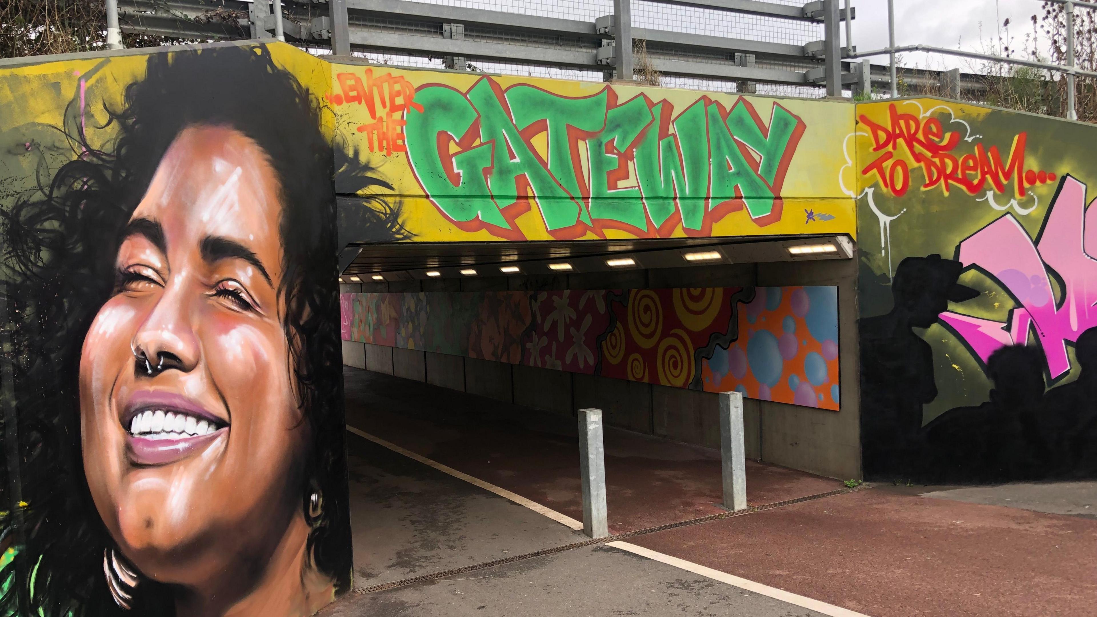 Colourful mural at the entrance to an underpass, a smiling woman has her eyes closed and appears to be in full sunshine on the left. "Enter the gateway" in bright green is above the 4 metre-wide entry point. "Dare to dream" in red with yellow border  is written on the right hand side. Further art works can be seen on the interior walls of the 6 metre-long concrete underpass